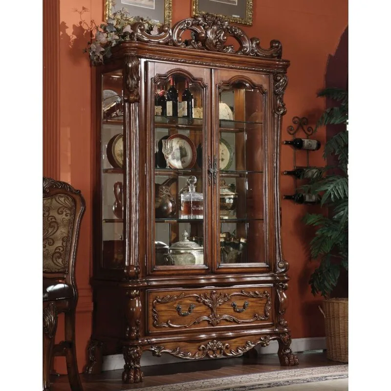 An ornate dark wood curio cabinet with intricate carvings and glass doors sits against a rich rust-colored wall displaying glass decanters and plates.