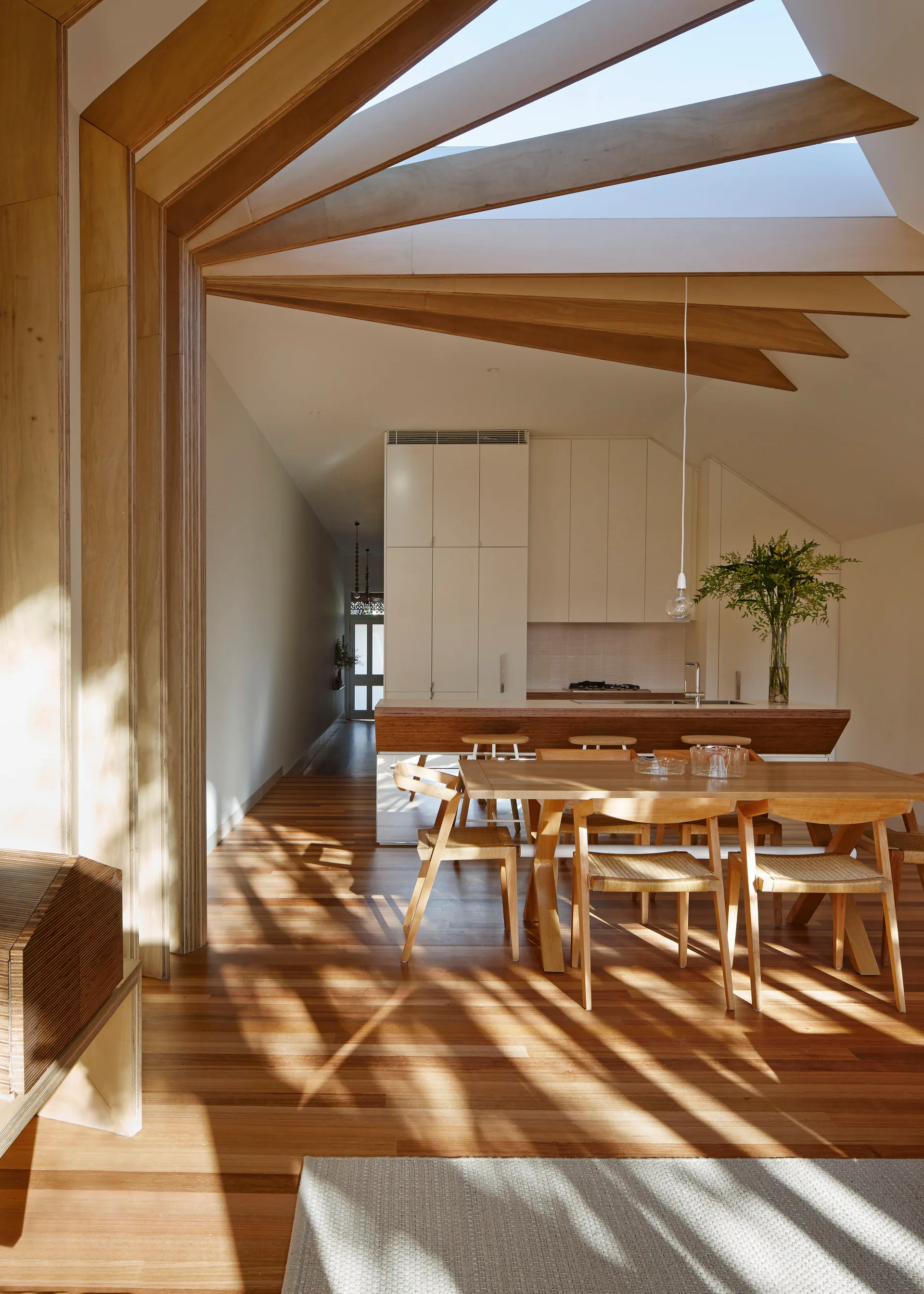 An open dining space features a wooden table, woven chairs, and a kitchen island beneath a skylight framed by angled timber rafters.