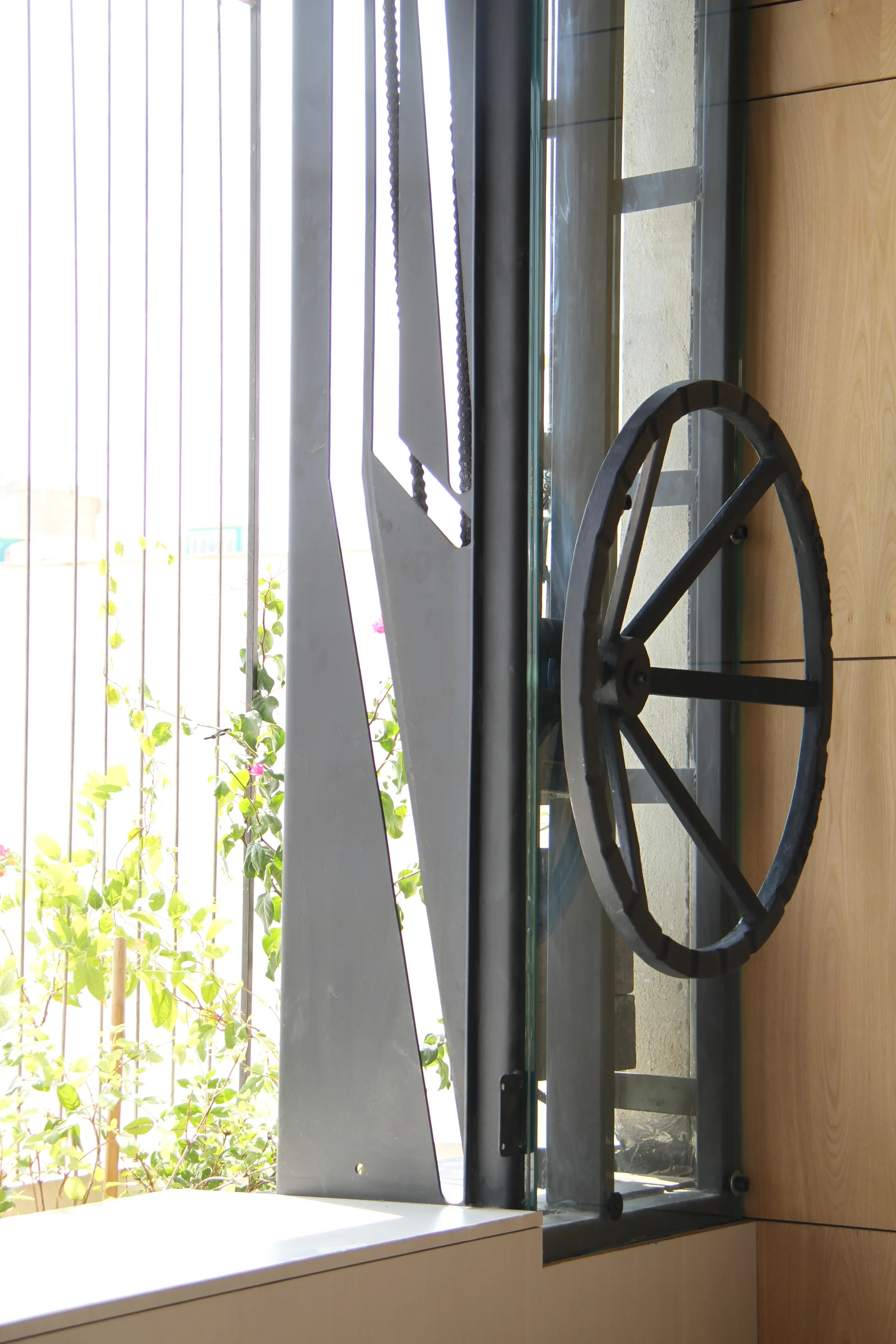 A close-up of a black metal industrial window mechanism with a large hand wheel set against light wood wall paneling.