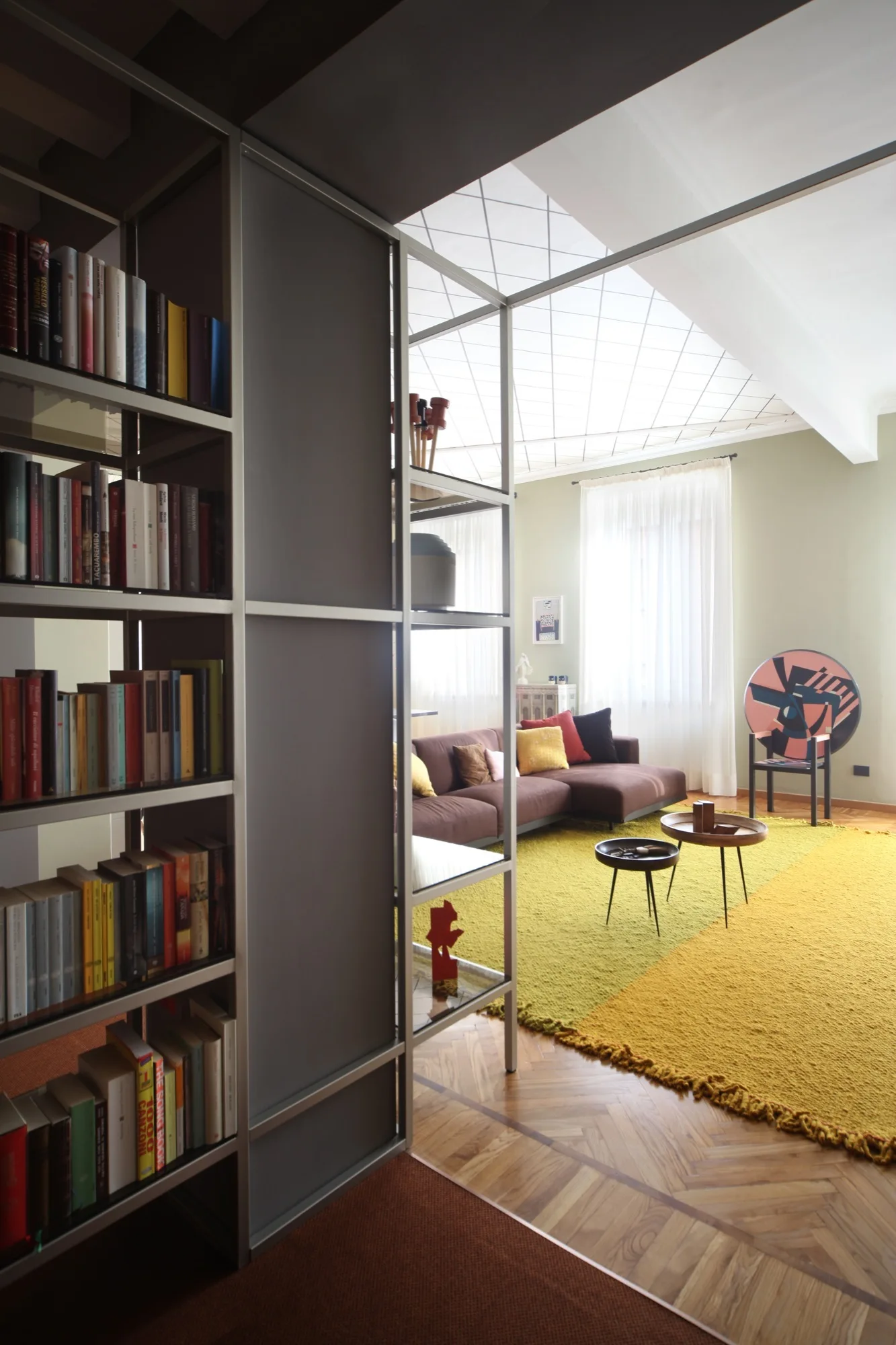 A modern living room featuring a tall metal bookshelf room divider, a brown sectional sofa, a textured yellow rug, and herringbone floors.