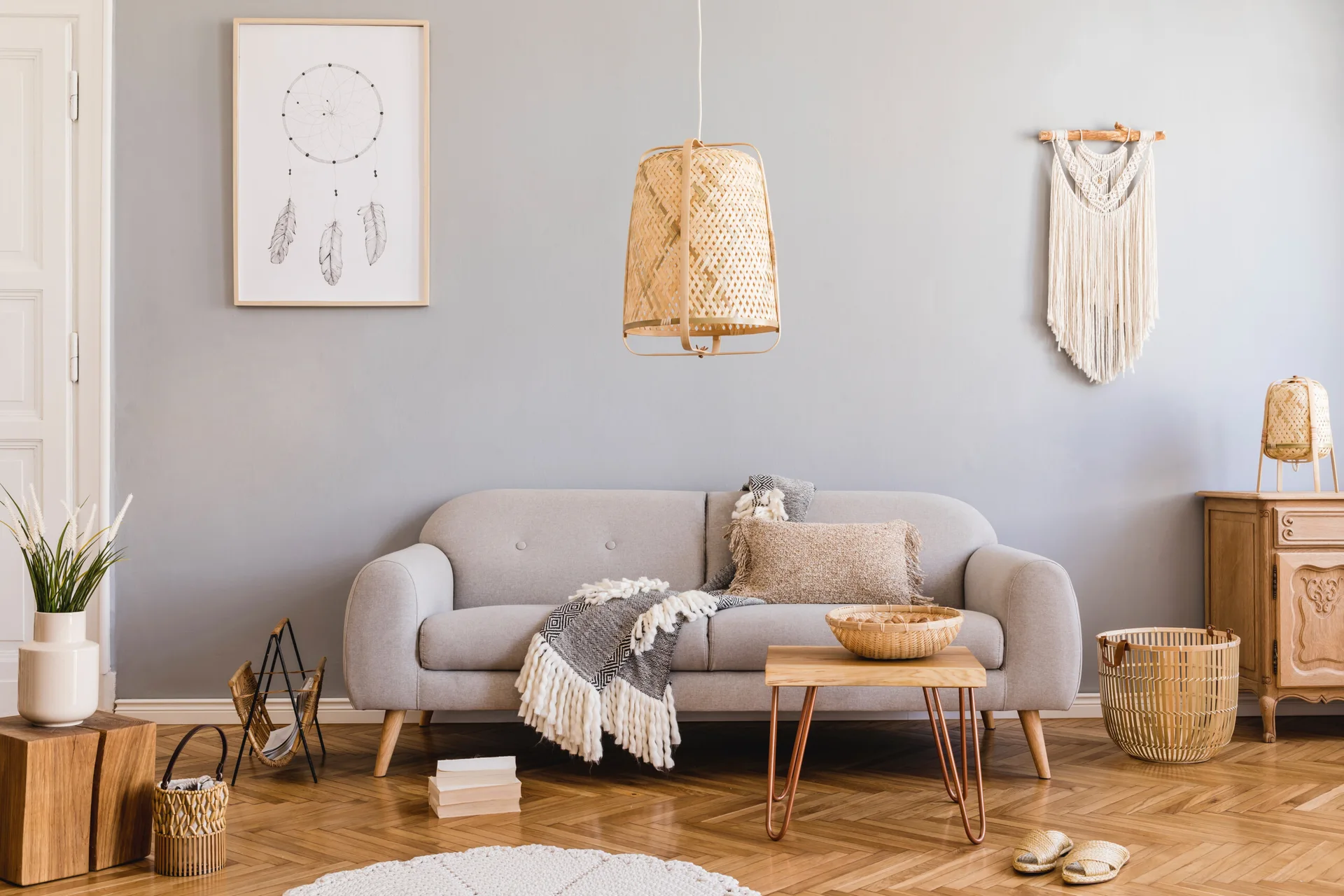 A grey sofa sits on a herringbone wood floor in a living room decorated with a woven pendant lamp, macrame wall hanging, and wooden furniture.