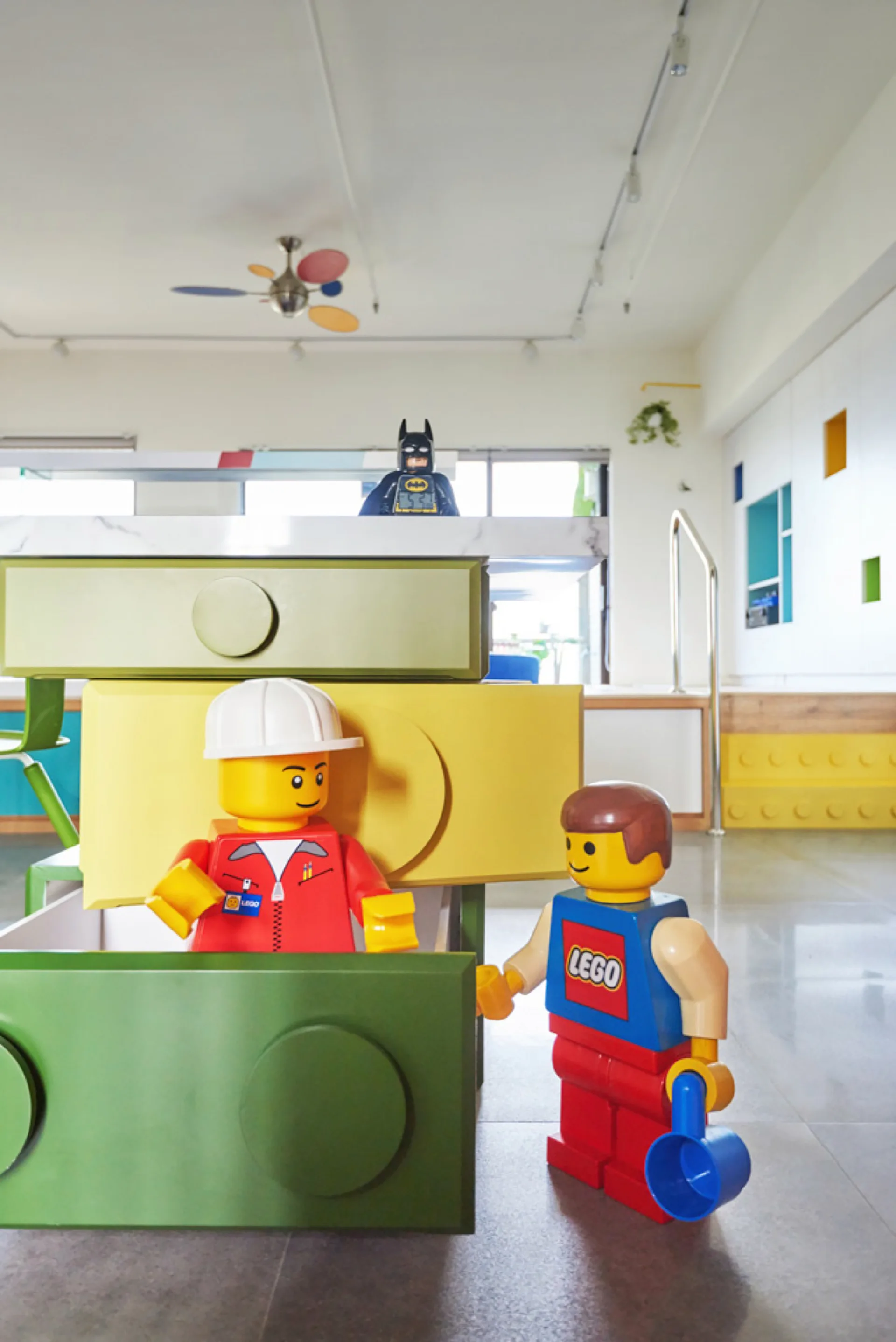 A playful kids room features storage cabinets shaped like green and yellow LEGO bricks with oversized figurines on a grey floor.