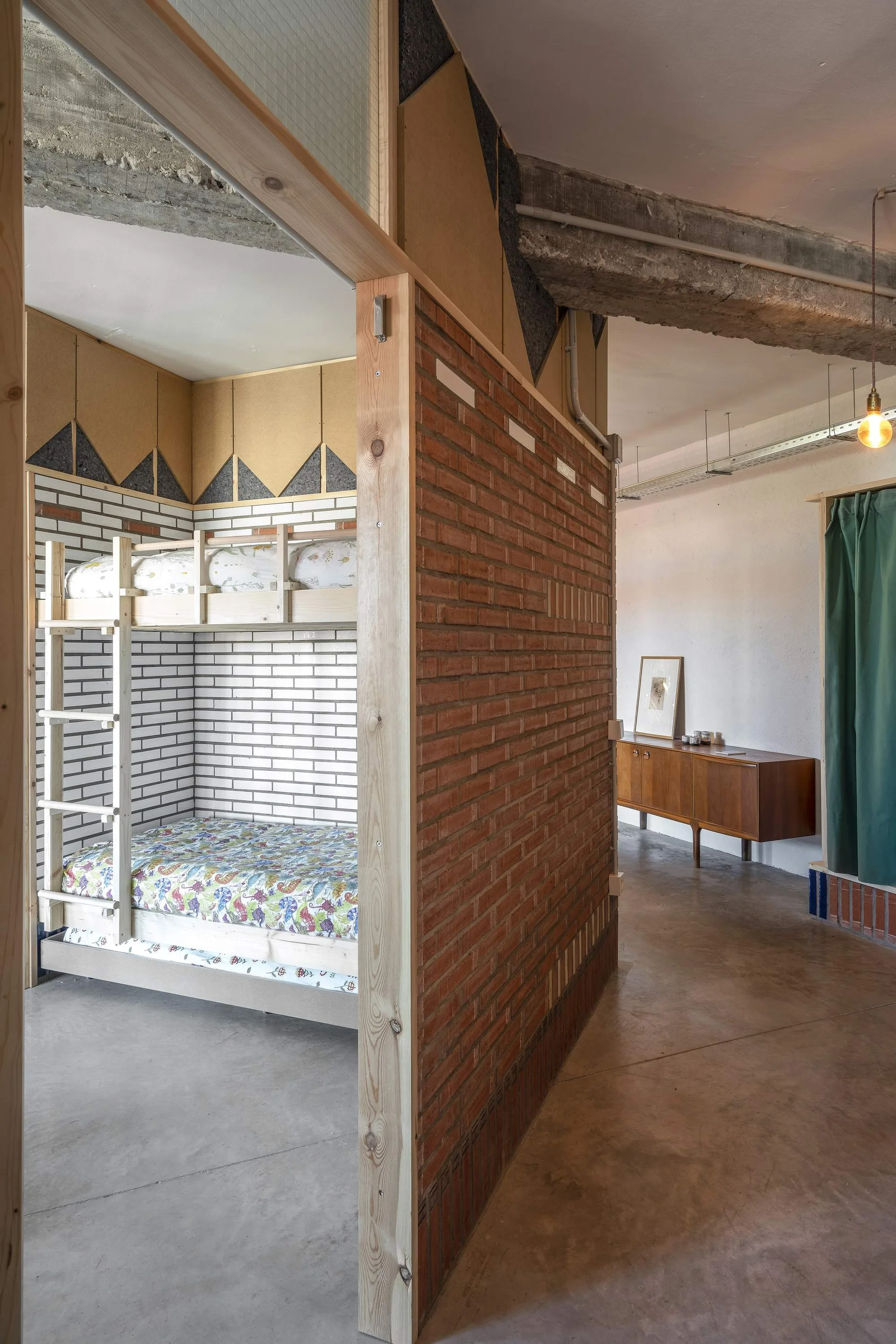 A light wooden bunk bed is nestled inside a cozy alcove behind a textured terracotta brick partition wall on a polished concrete floor.