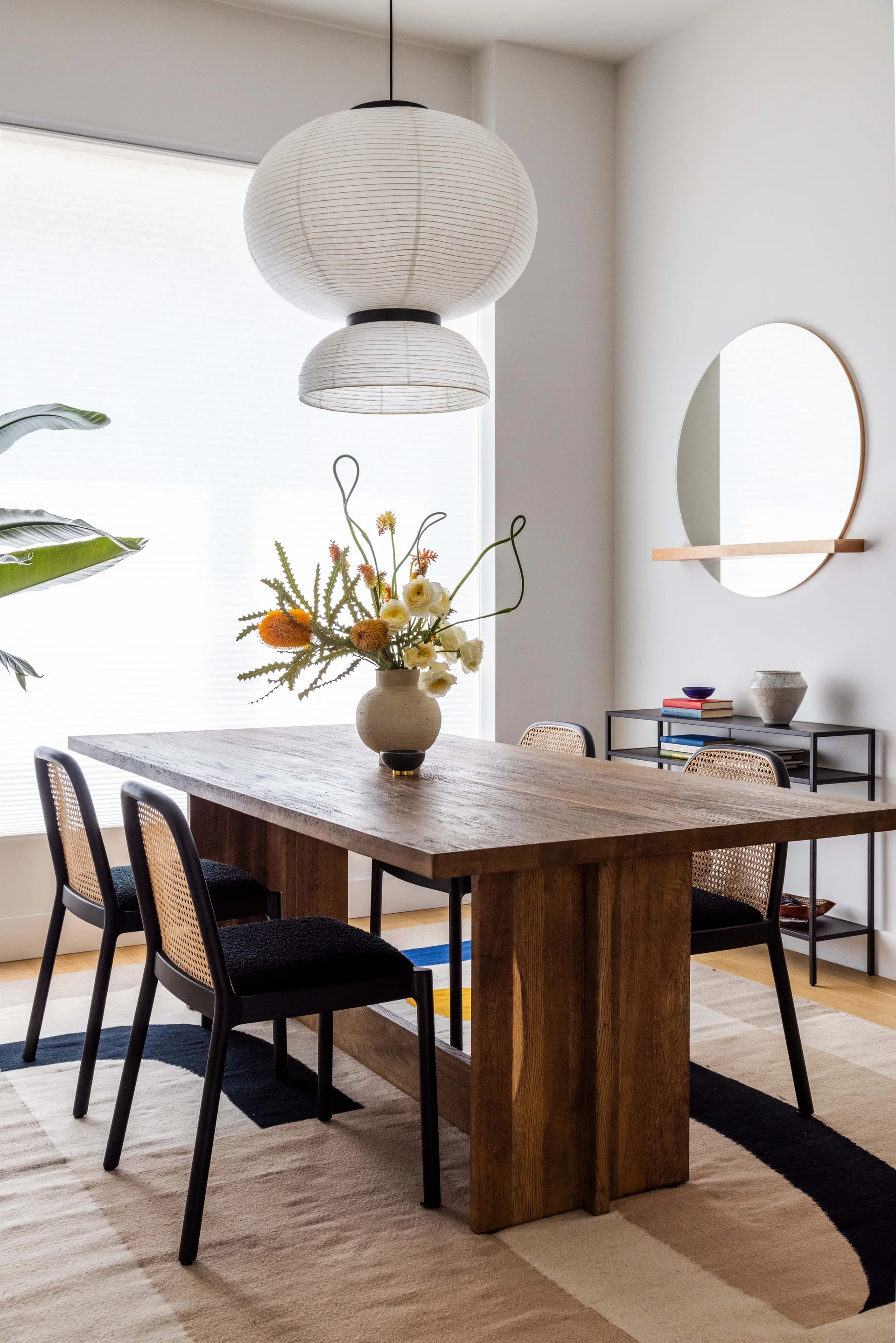 A light wood dining table paired with black cane chairs sits on a patterned rug under a large tiered paper pendant light.