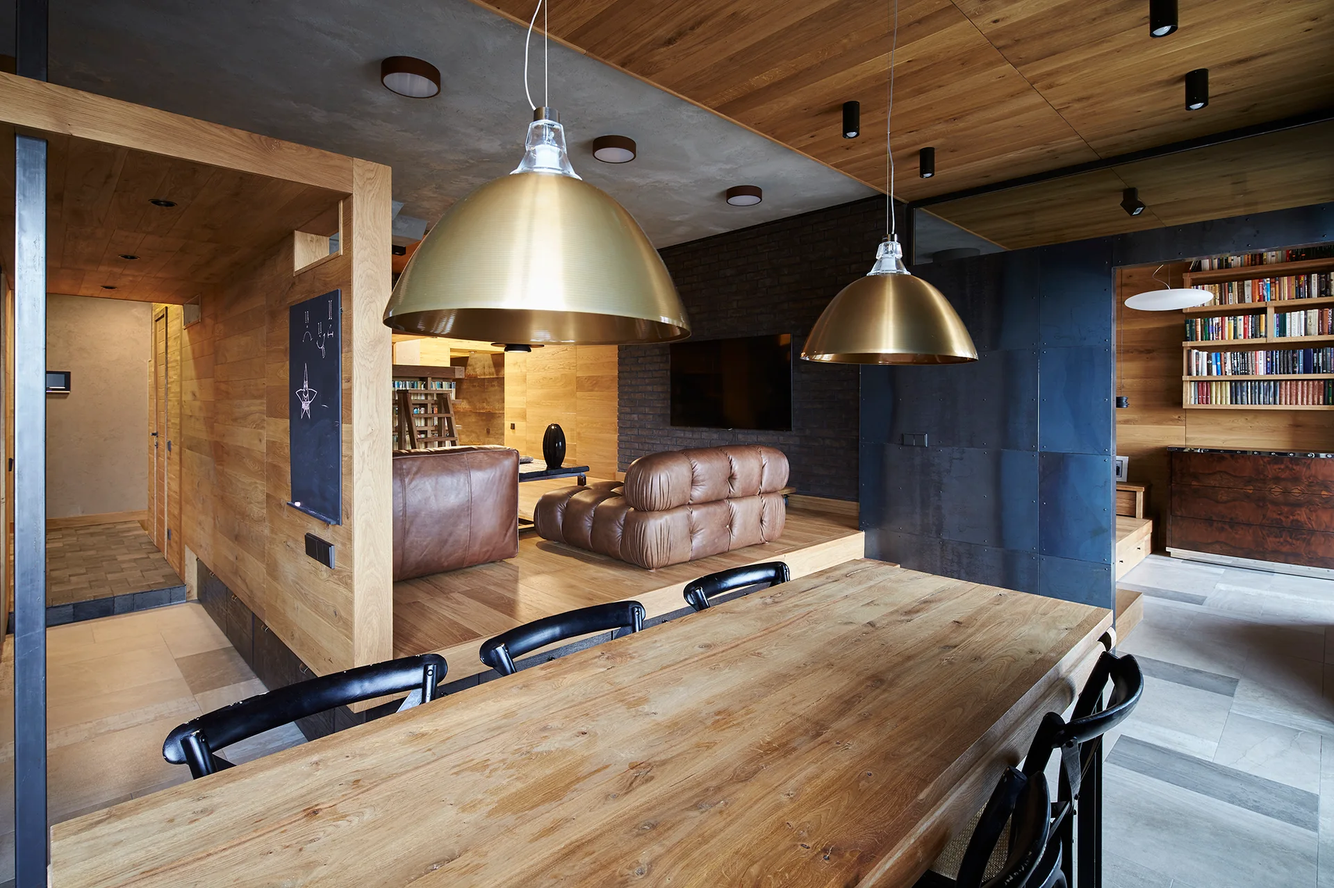 An industrial living and dining area featuring a raised wood platform, tufted leather sofas, a wooden dining table, and brass dome pendant lights.