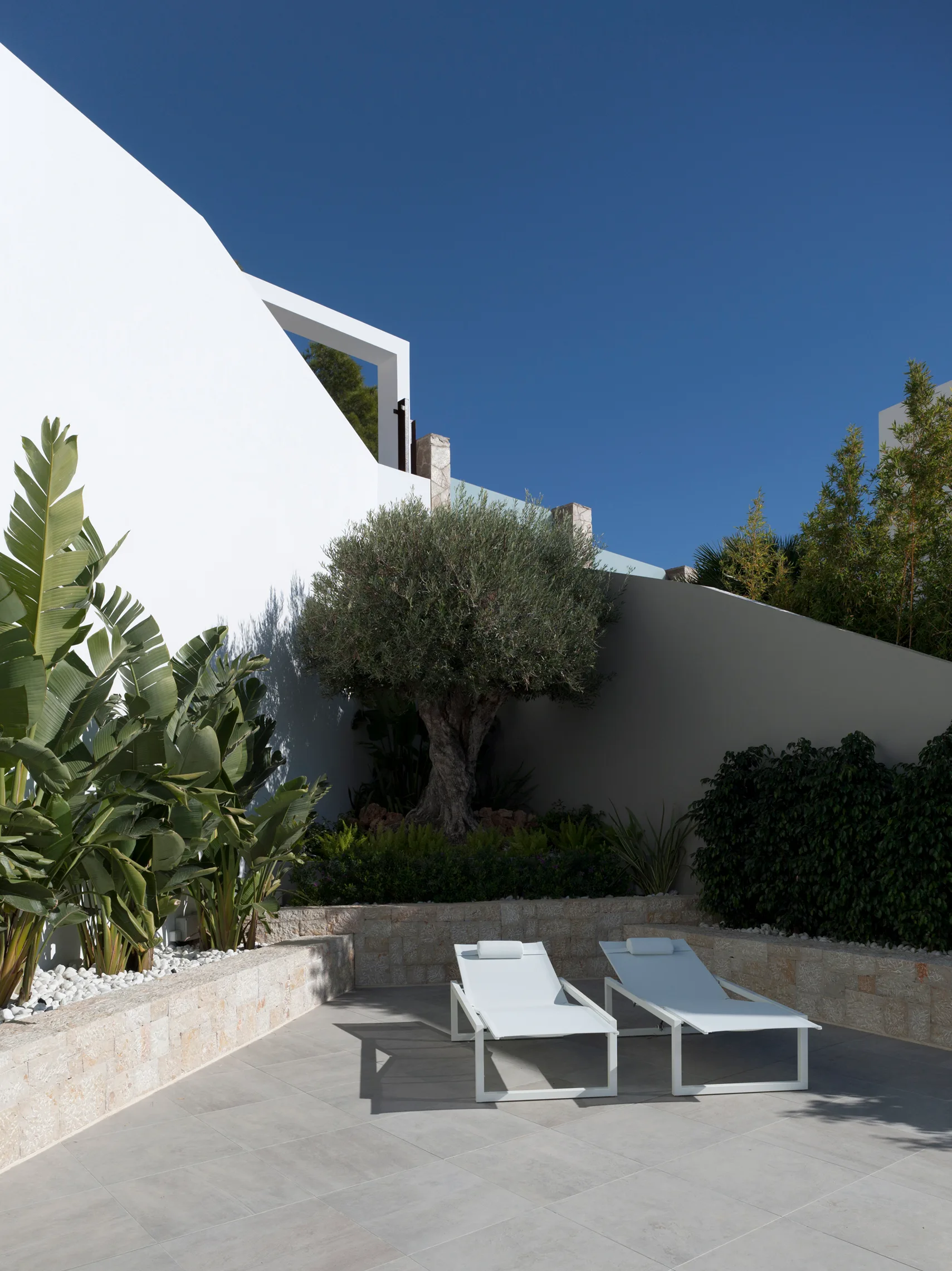 Two minimalist white sun loungers rest on a paved gray patio beside a stone retaining wall with lush green plants and a white architectural wall.