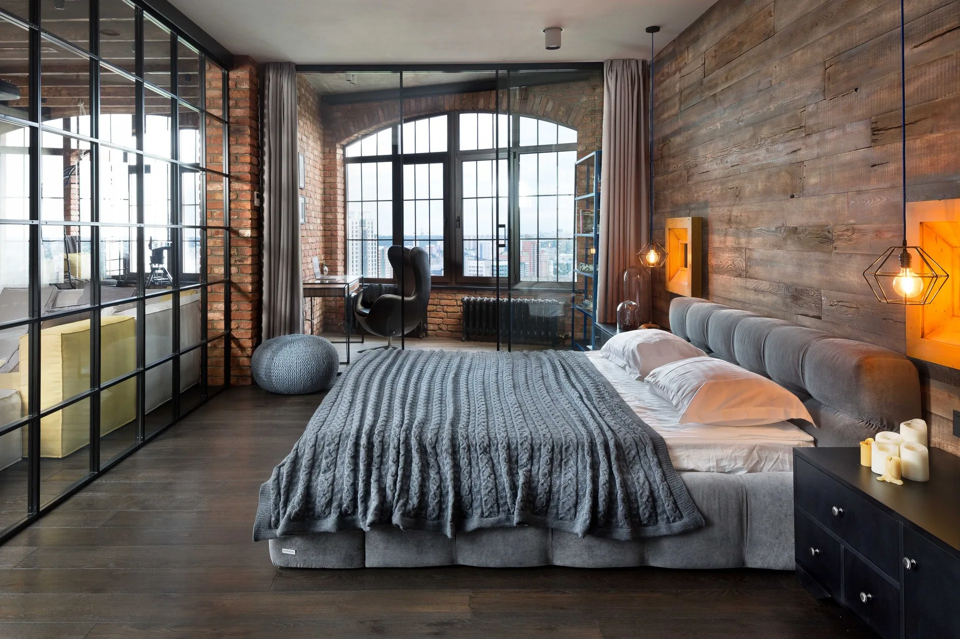 An industrial bedroom featuring a grey upholstered bed against a wood-paneled wall, alongside a glass-partitioned workspace with arched windows.