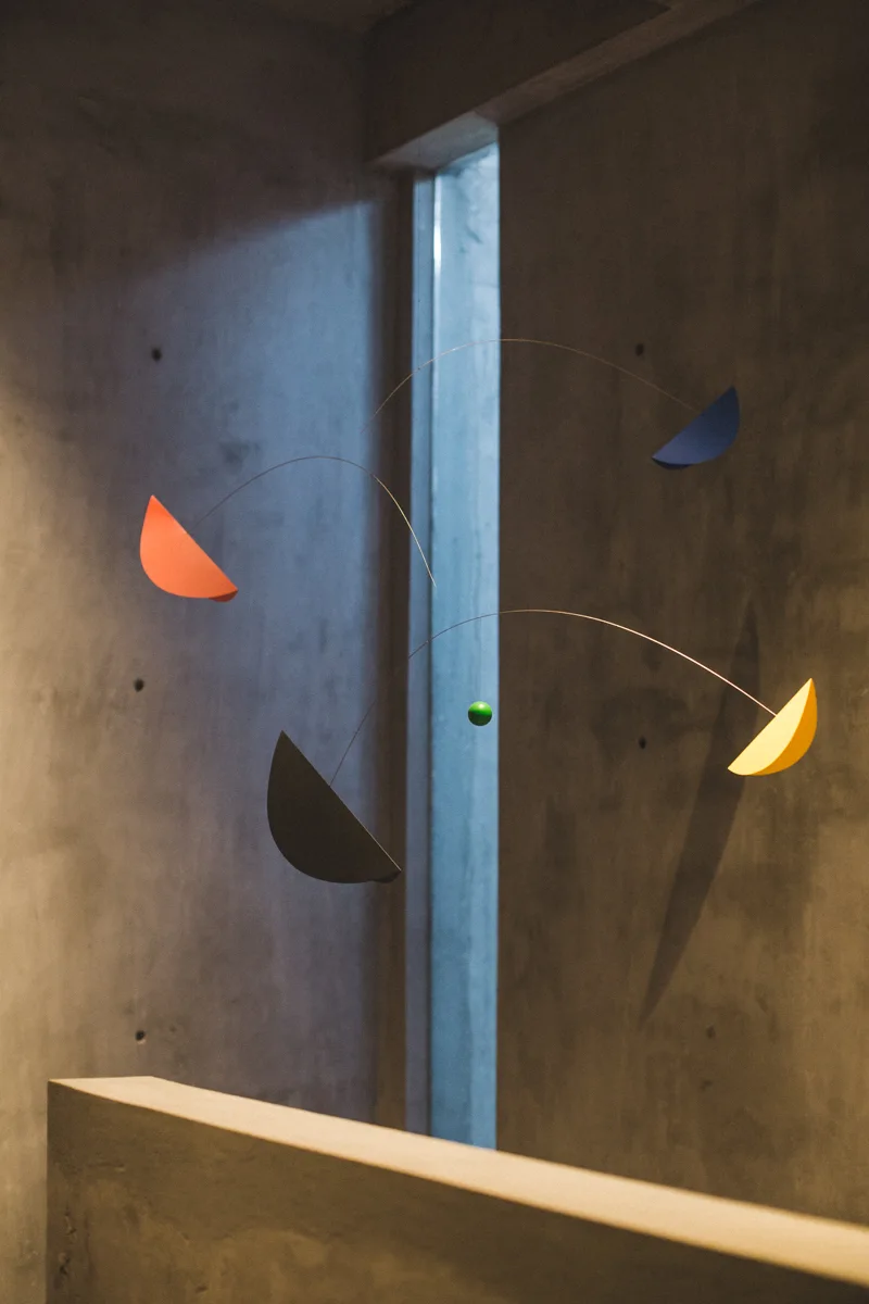 A colorful kinetic mobile with yellow, orange, and blue shapes hangs in front of a textured gray concrete wall next to a narrow window.