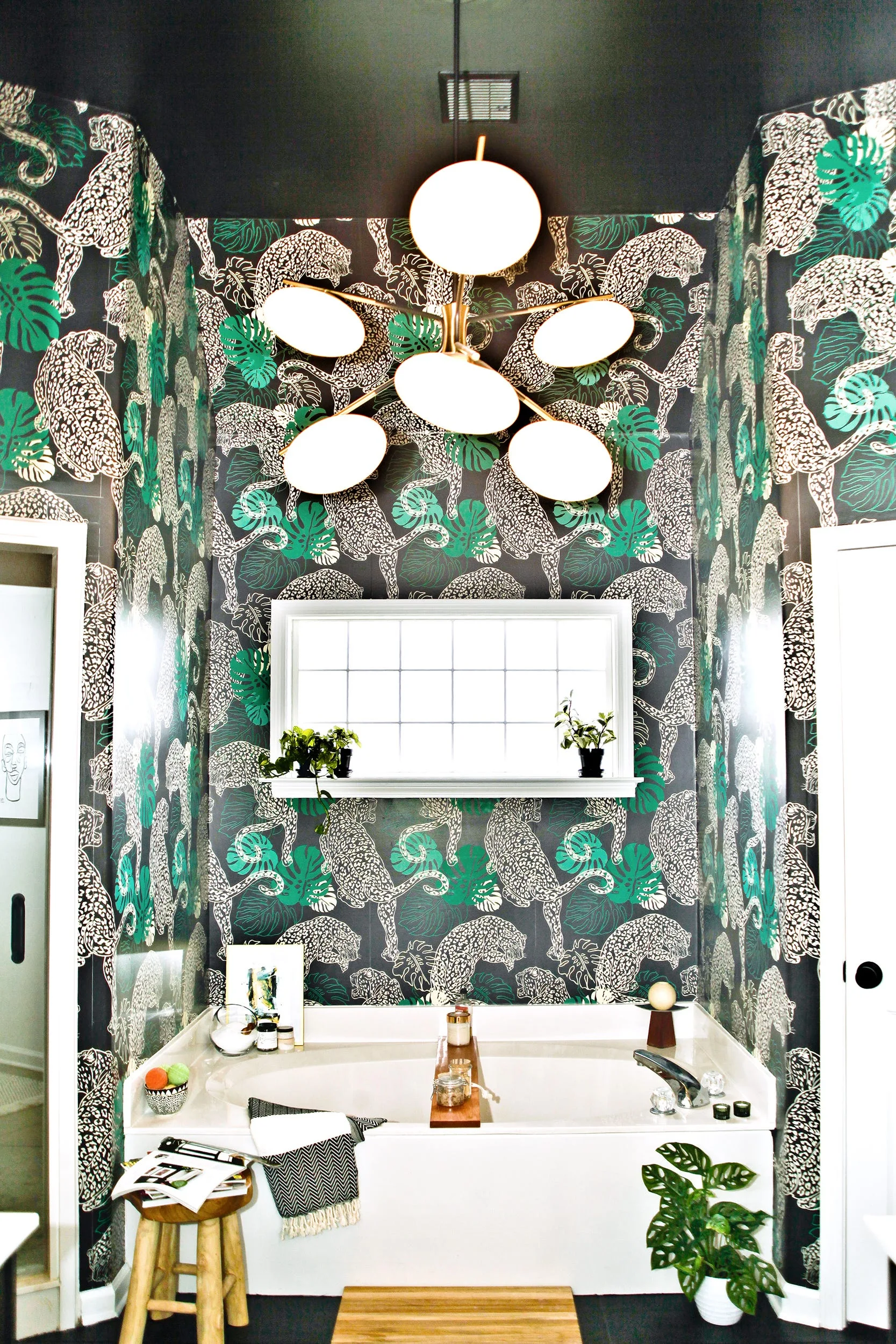 A white bathtub is set against black, white, and green leopard-print wallpaper under a dark ceiling with a modern brass chandelier.