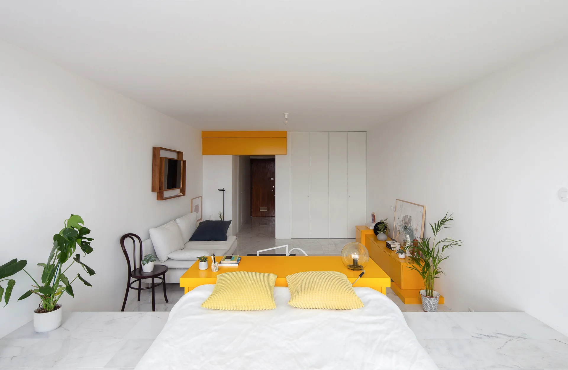 A minimalist white studio apartment featuring a bed with yellow pillows, a matching yellow console table, and a small sofa.