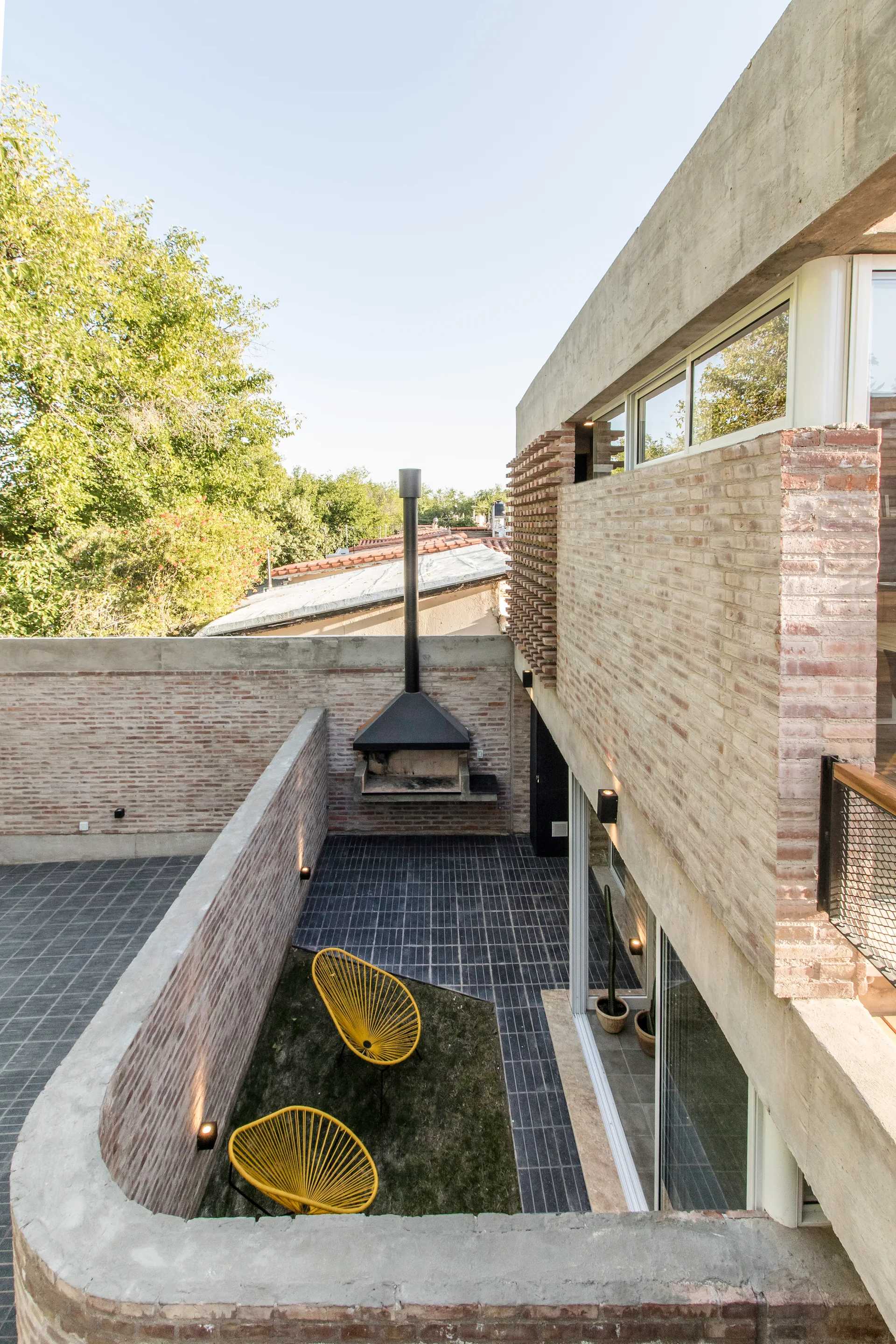 An outdoor brick patio features dark tiled floors, a black metal fireplace, and two yellow woven chairs situated on a small grass area.