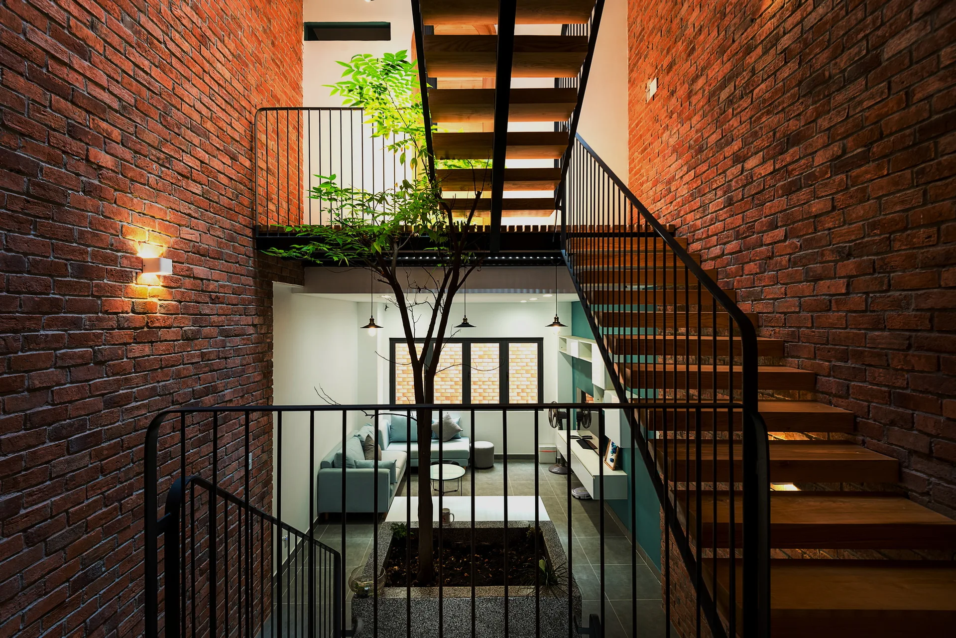 An open wood and metal staircase wraps around a tall brick atrium featuring a living tree planted in a stone container on the lower level.
