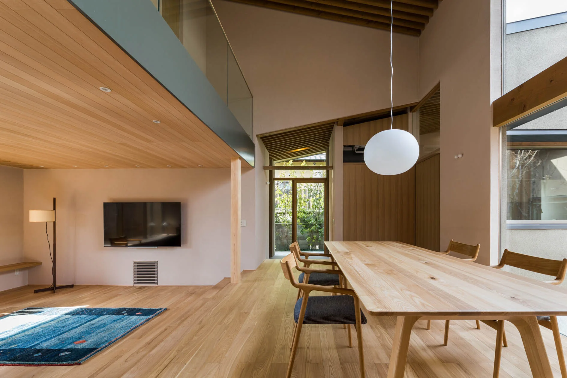 A minimalist dining room features a light wood table and chairs beneath a modern white globe pendant light and a glass-railed mezzanine.