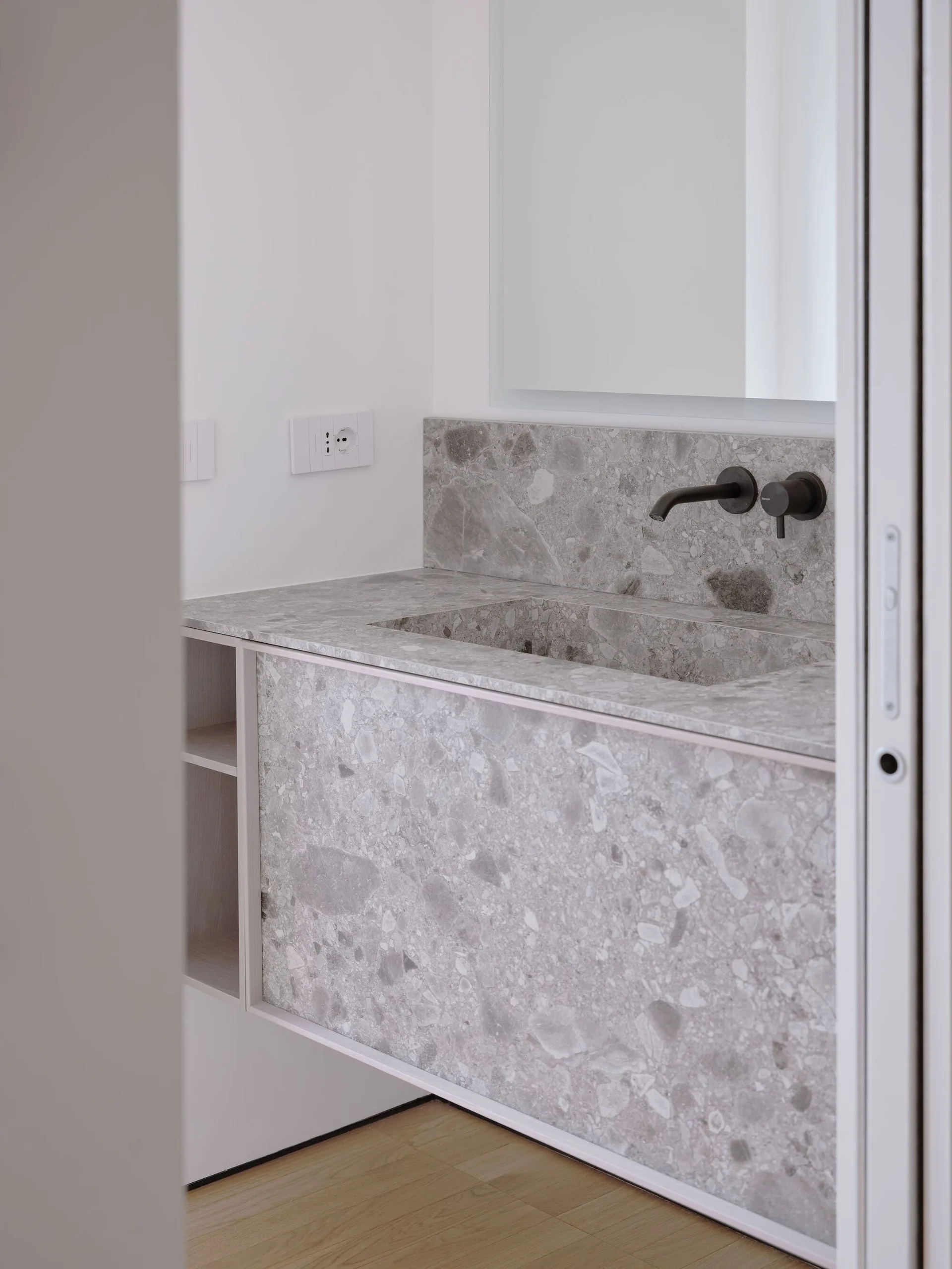 A modern bathroom vanity made from grey speckled stone features an integrated sink basin and a matte black wall-mounted faucet below a simple mirror.