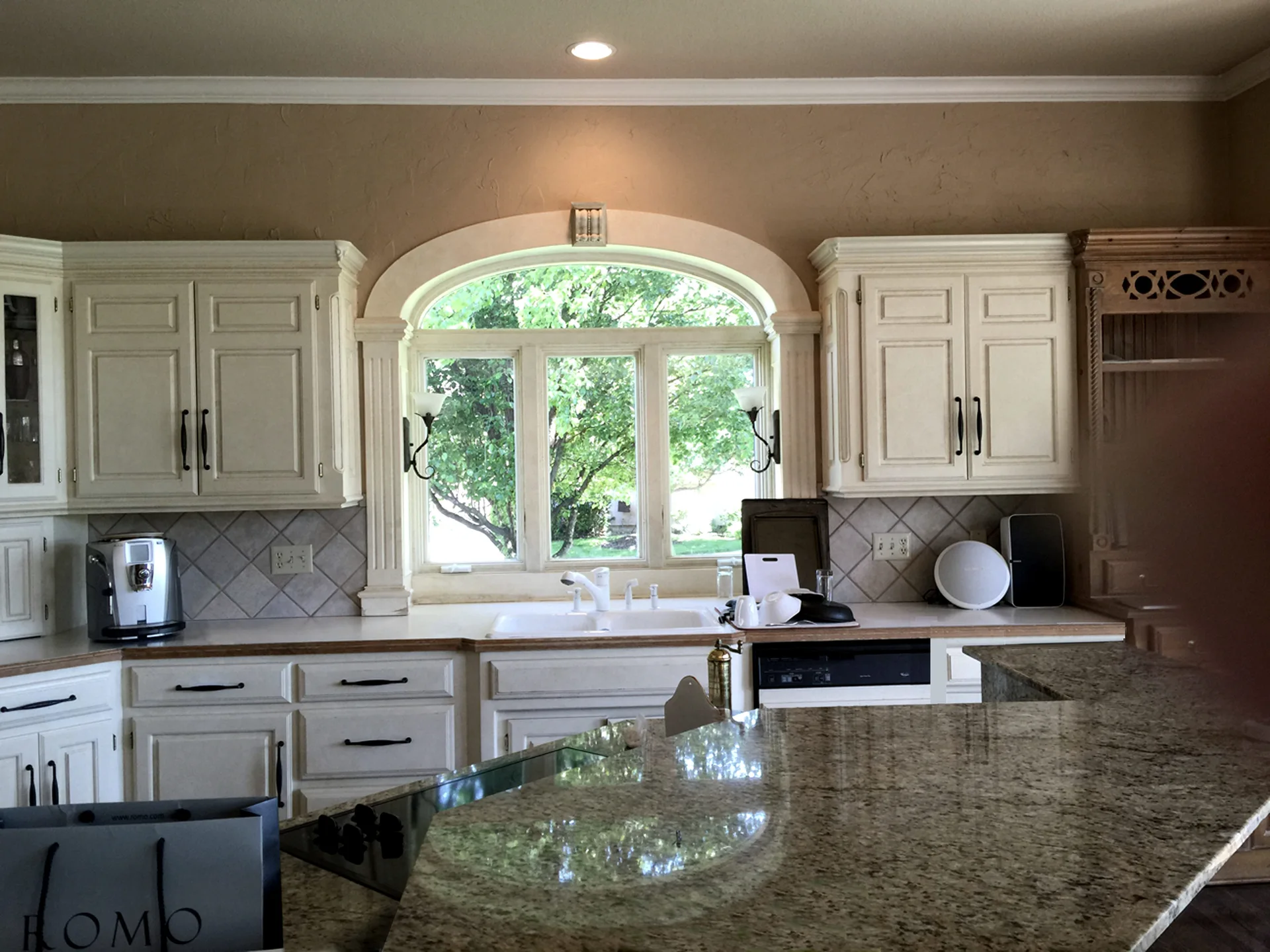 A kitchen with cream cabinets, a double sink beneath an arched window, and a speckled granite island in the foreground.