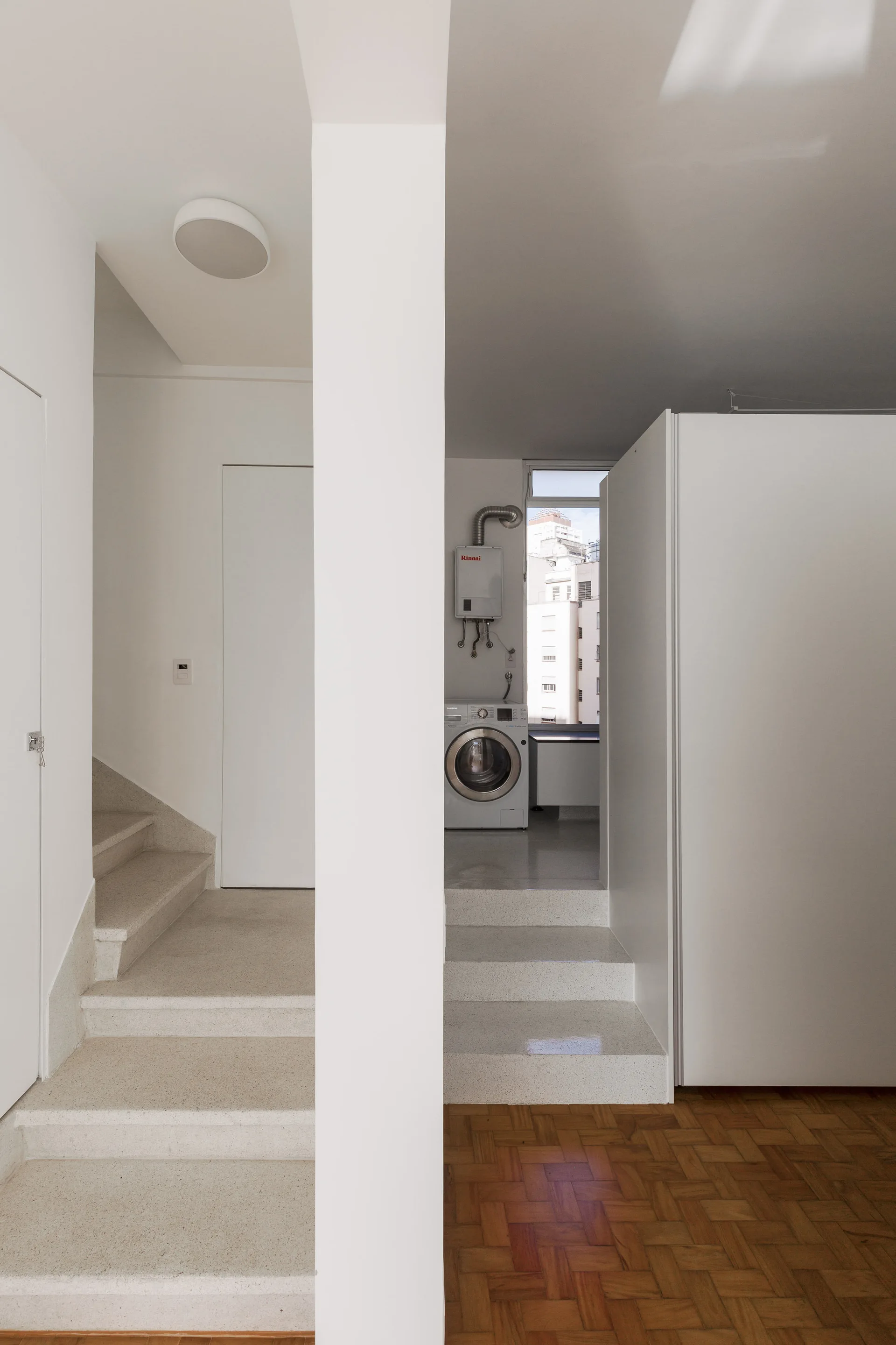 White minimalist interior with stone steps leading up to a raised laundry area next to a central pillar and parquet wood flooring.