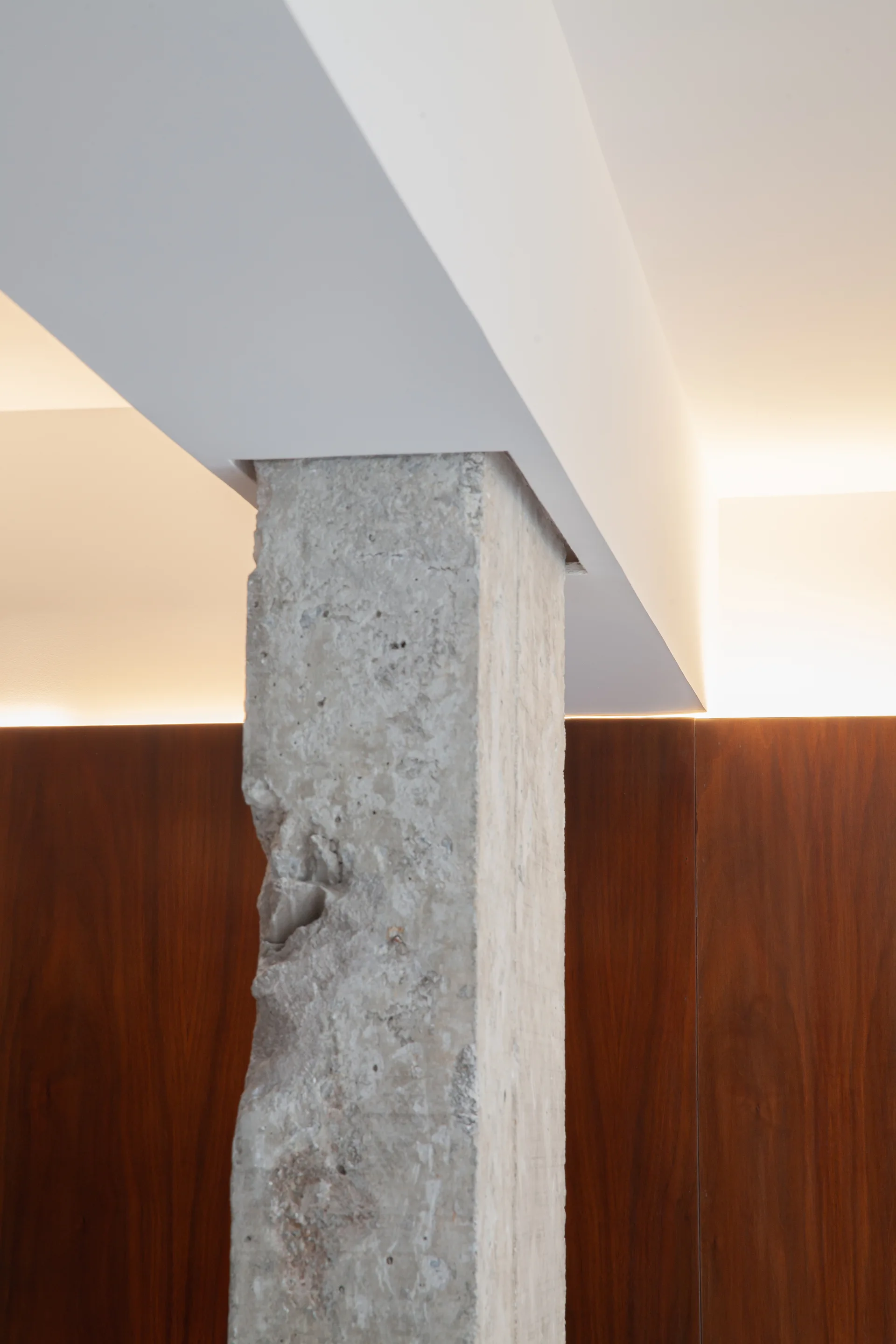 A rough, textured concrete column stands in front of dark wood wall paneling beneath a smooth white ceiling beam with integrated cove lighting.