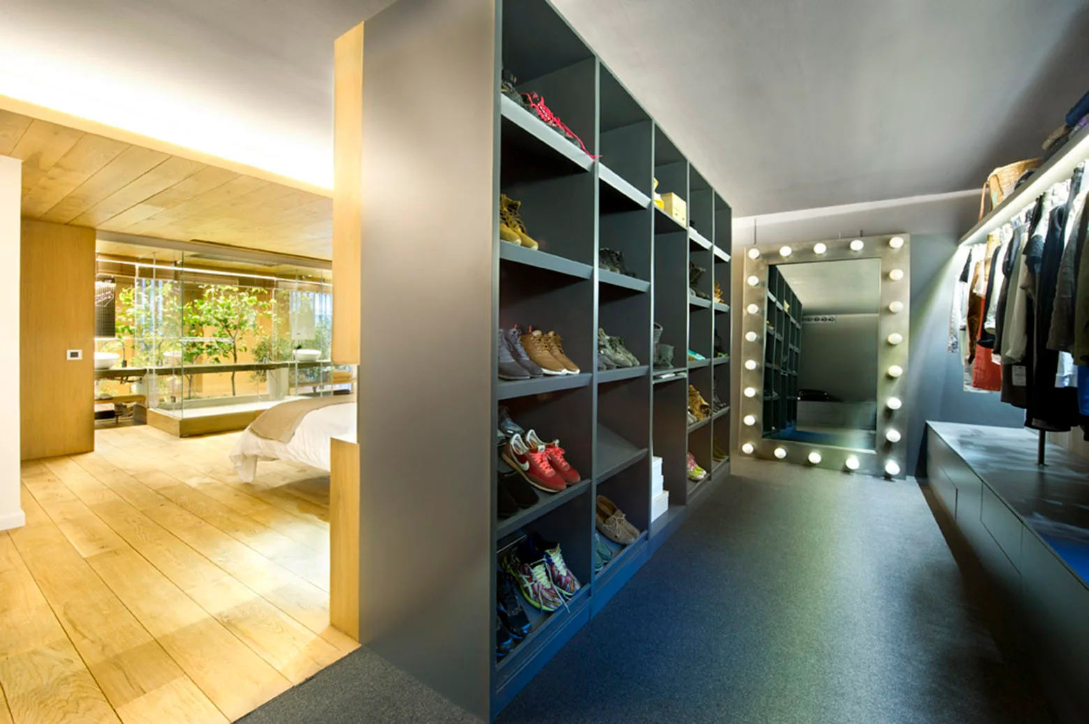 A modern walk-in closet featuring a large dark grey shoe shelving unit, a lighted vanity mirror, and an opening to a wood-floored bedroom.