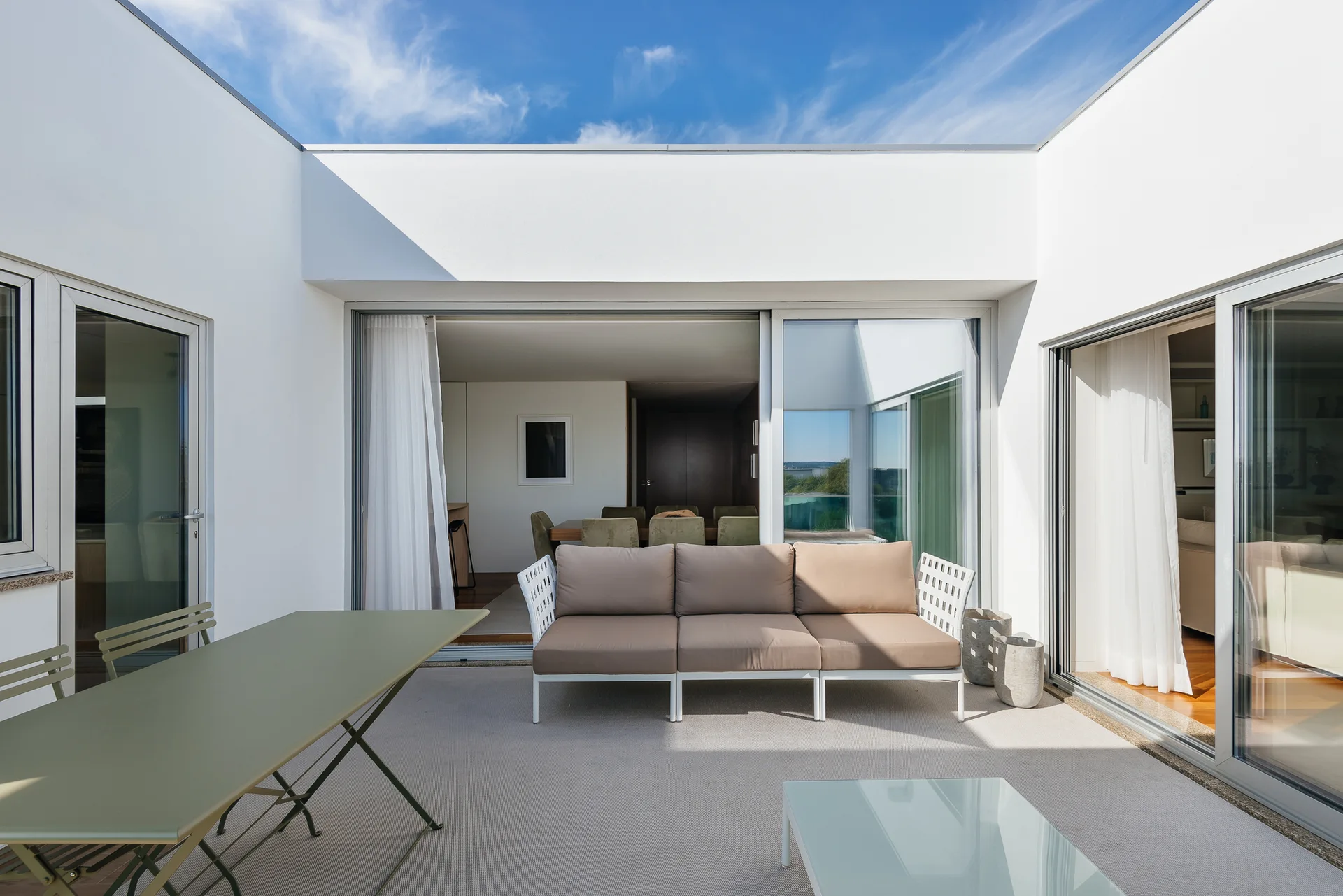 A sunny white stucco courtyard patio features a modular beige sofa and a light green table next to sliding glass doors showing the indoor dining room.