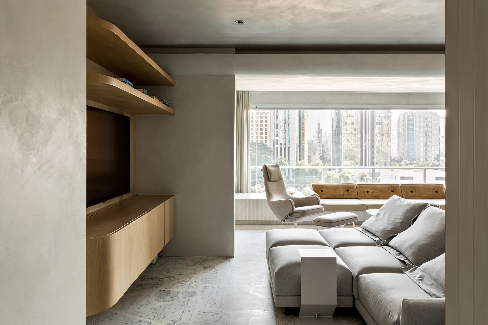 A minimalist living room features a grey modular sectional sofa, veined marble floor tiles, and custom wood cabinets with curved edges.