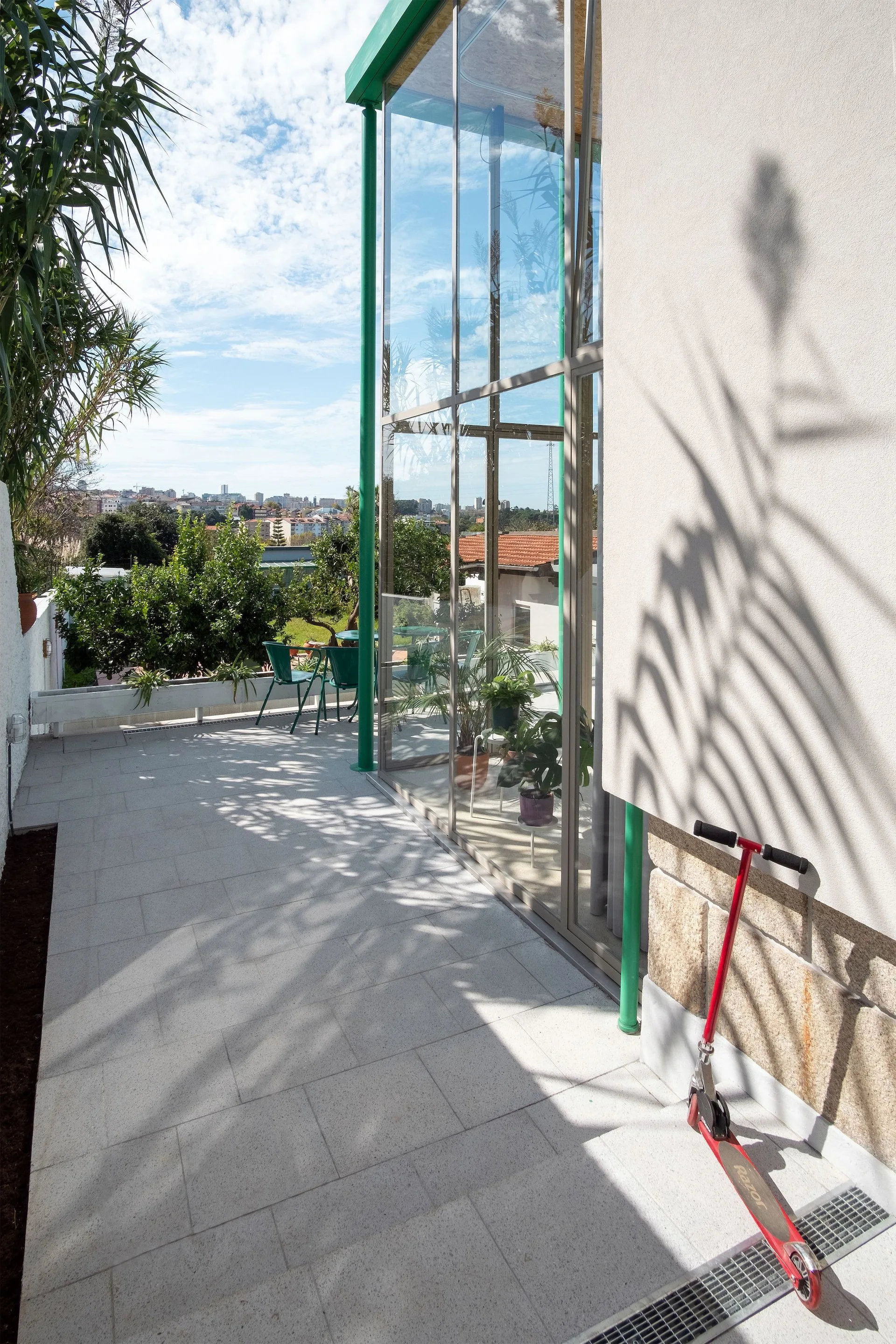 A paved outdoor patio features a tall glass facade, green metal patio furniture, potted plants, and a red scooter resting against a white plaster wall.