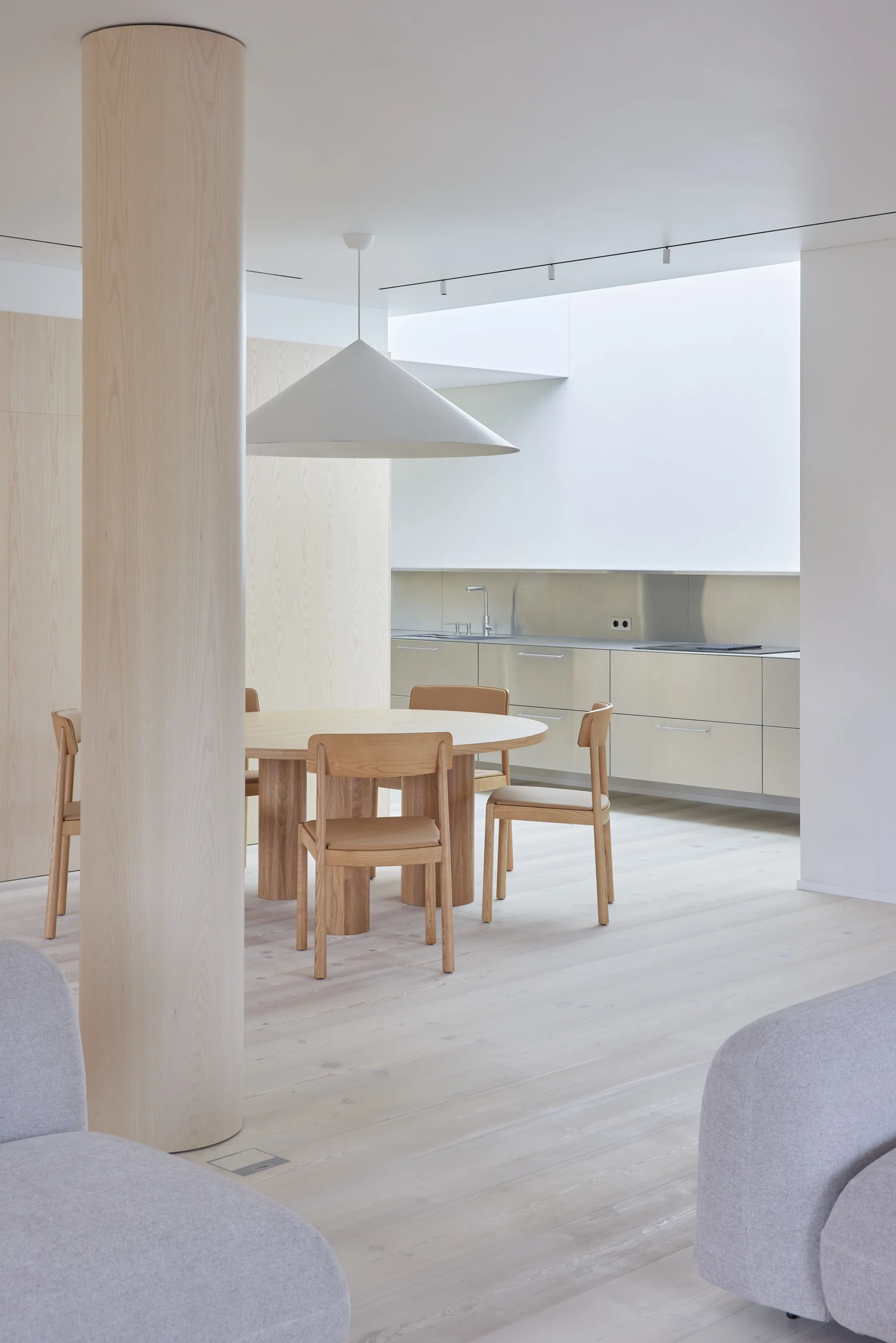 A minimalist dining room features a round light wood table and chairs next to a large cylindrical wood column and a sleek stainless steel kitchen.