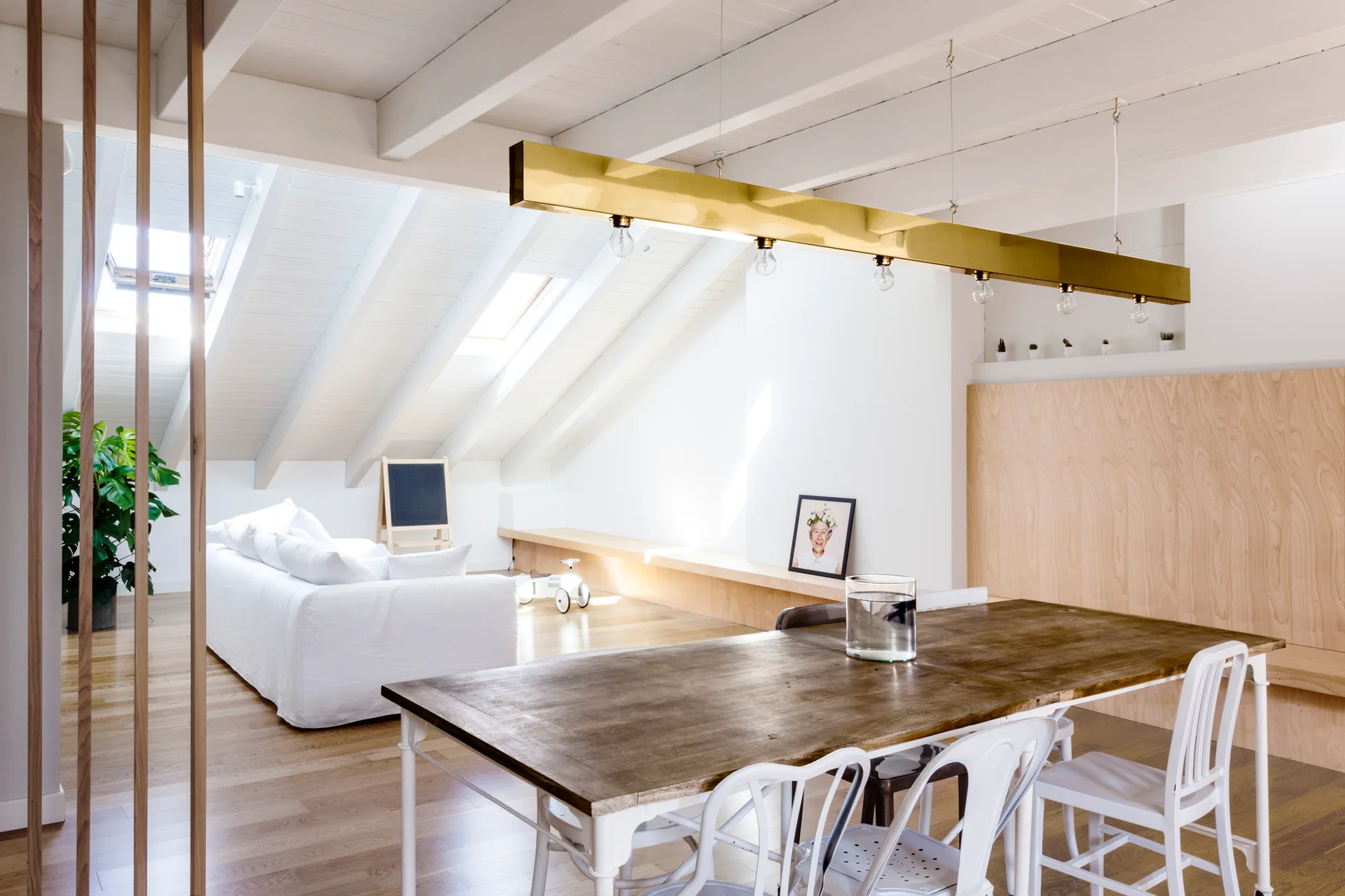 A bright attic dining room featuring a wooden table with white metal chairs under a long brass pendant light, beneath a sloped white ceiling.