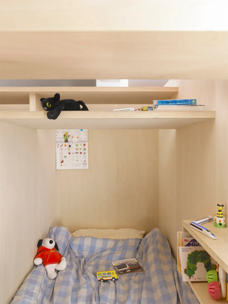 A cozy child's sleeping nook enclosed in light-toned wood paneling features built-in shelving, blue plaid bedding, and small toys.