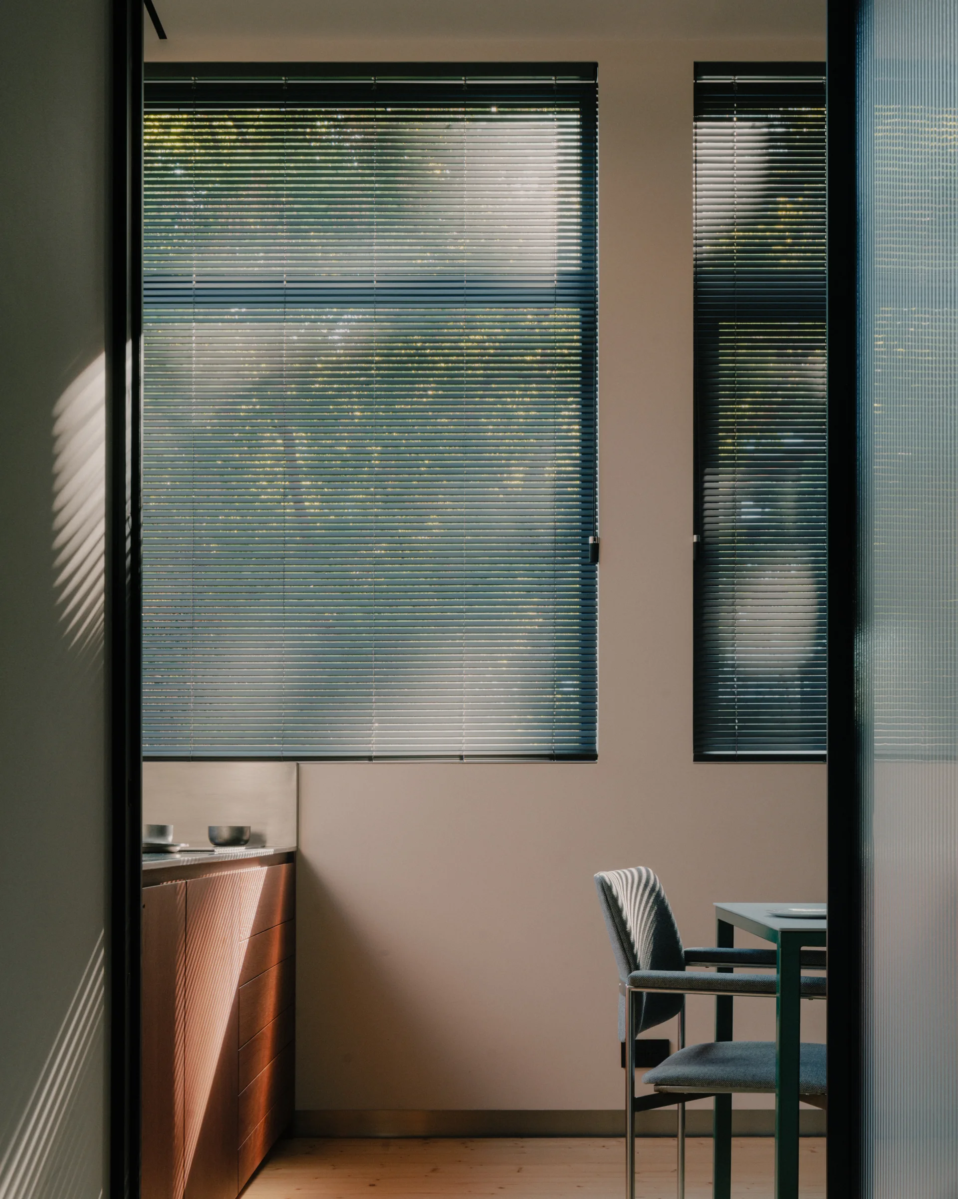 A view through a doorway into a dining area with dark slatted window blinds, an upholstered chair, and wood cabinetry.