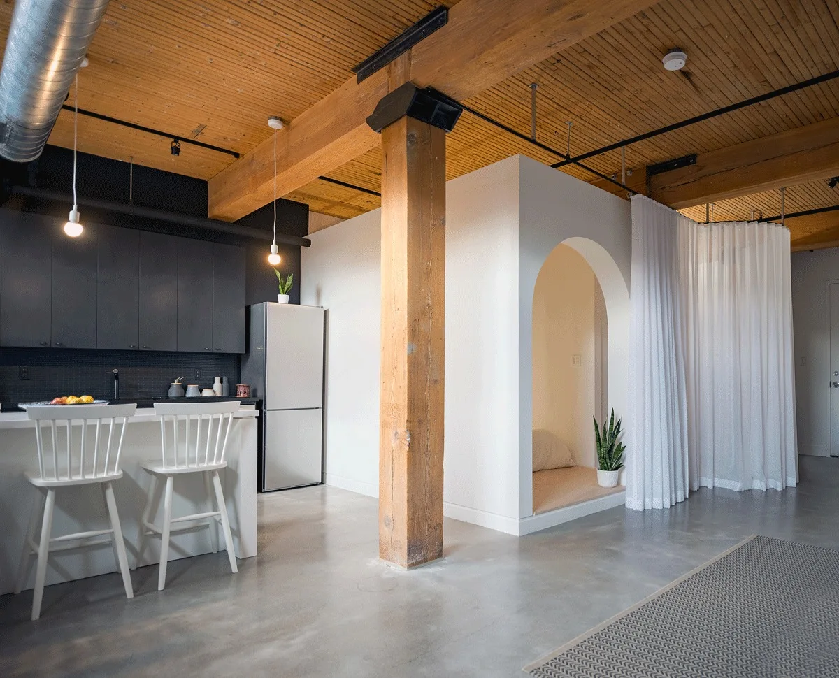 An industrial loft kitchen with black cabinets and a white island next to an arched white alcove and exposed wood ceiling.