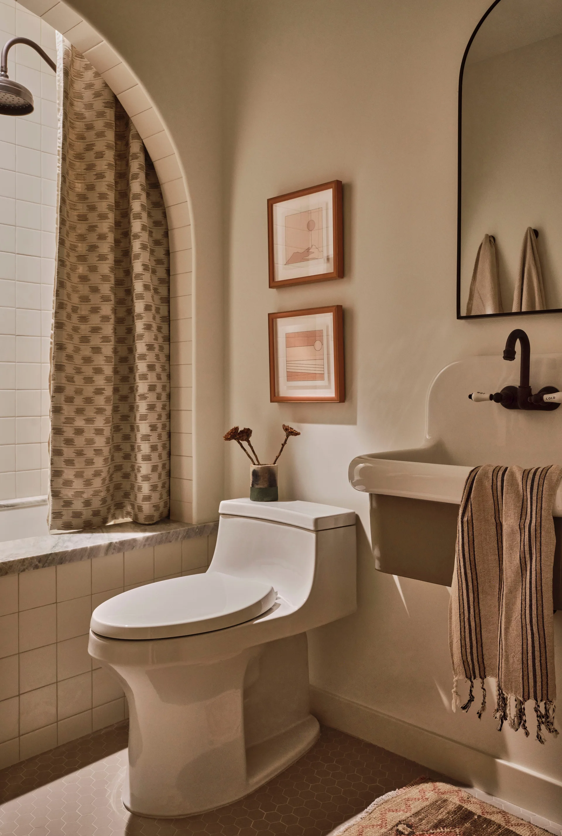 A warm beige bathroom features a white ceramic toilet, a wall-mounted utility sink with black hardware, and an arched shower entryway with a curtain.