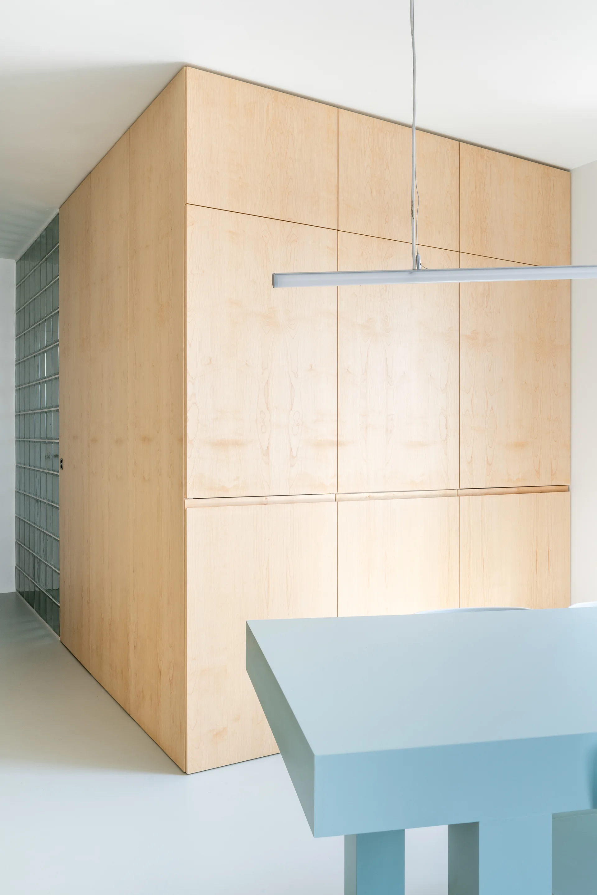 A modern room featuring a large floor-to-ceiling light wood storage cabinet, a matte light blue desk, and a translucent glass block wall.