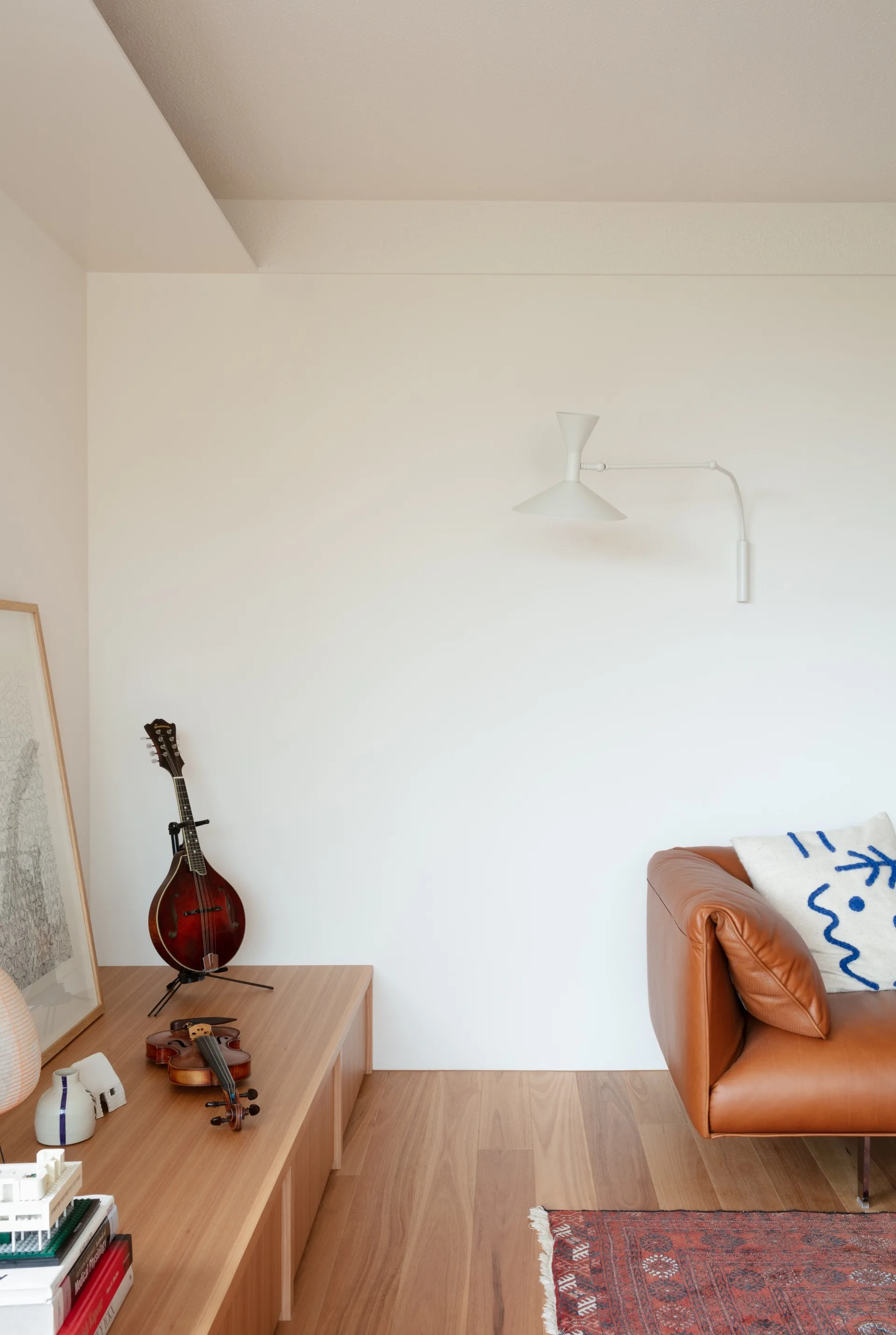 A minimalist living room corner features a brown leather sofa, a low wood console displaying a violin and mandolin, and a white swing-arm wall sconce.