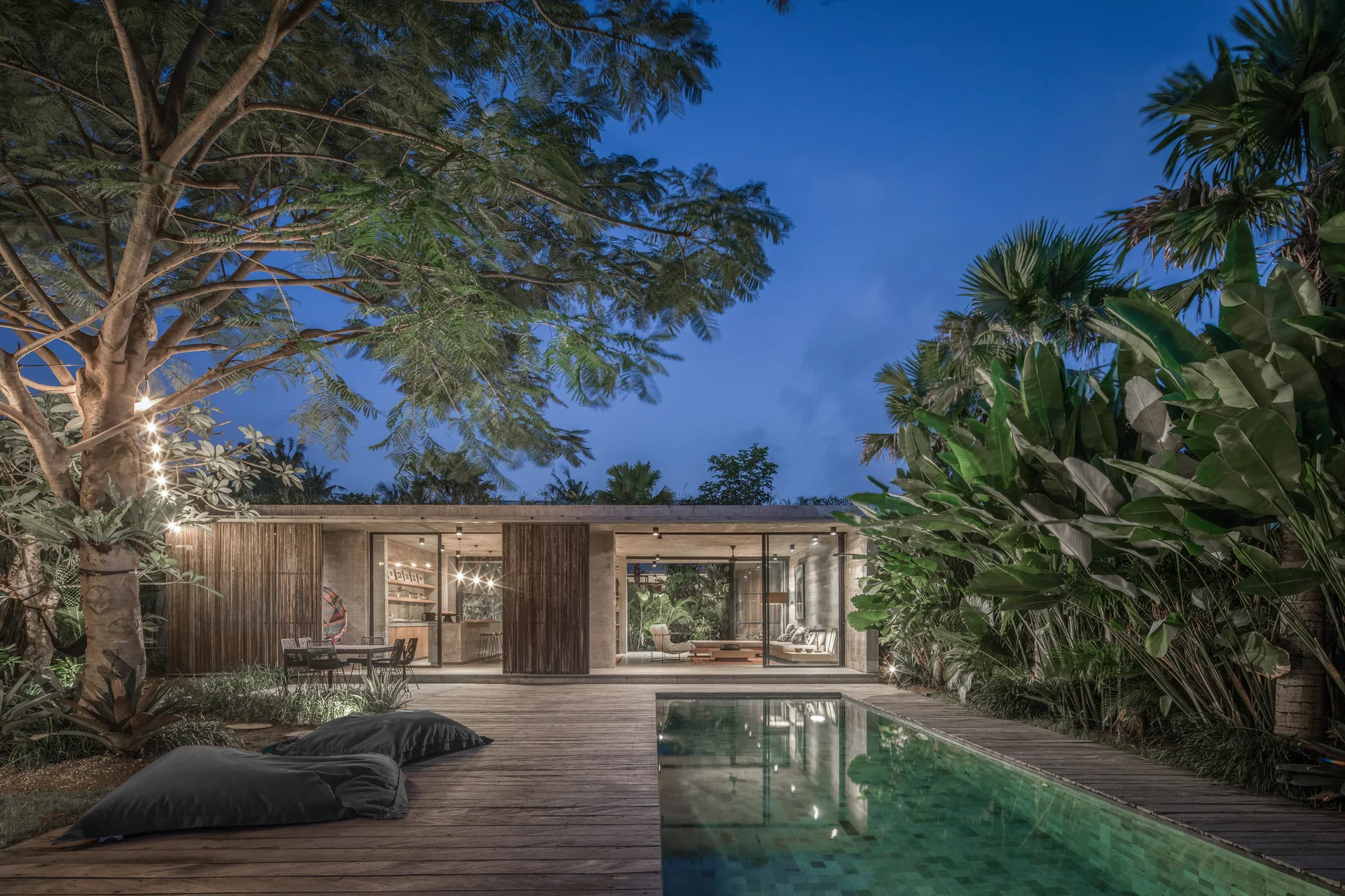 A rectangular swimming pool set in a wooden deck next to an open concrete villa with glass walls, surrounded by dense tropical foliage at twilight.