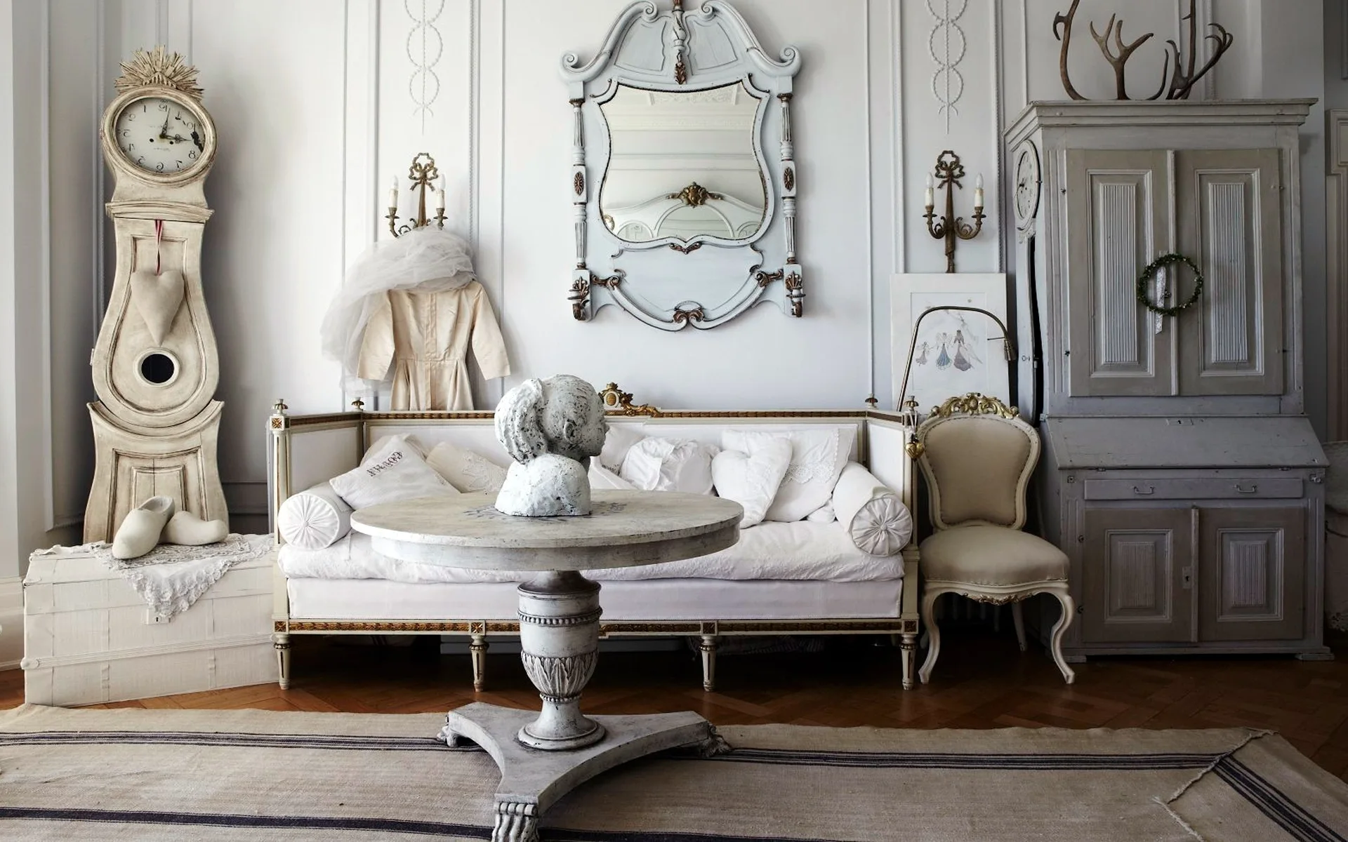 A light gray living room features a distressed white sofa, a circular pedestal table with a bust, an ornate mirror, and a vintage grandfather clock.
