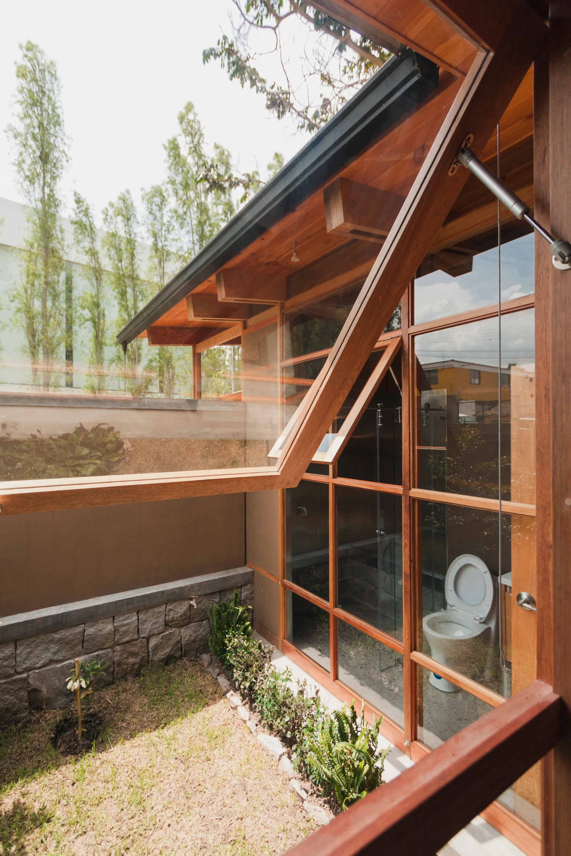 A wood-framed glass window wall looks into a modern bathroom with a visible toilet, showing exposed ceiling beams and an outdoor garden path.