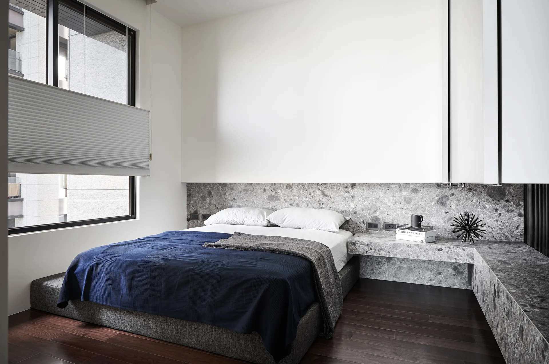 A low platform bed with a deep blue blanket sits on dark wood floors next to a floating grey speckled stone nightstand and a large window.