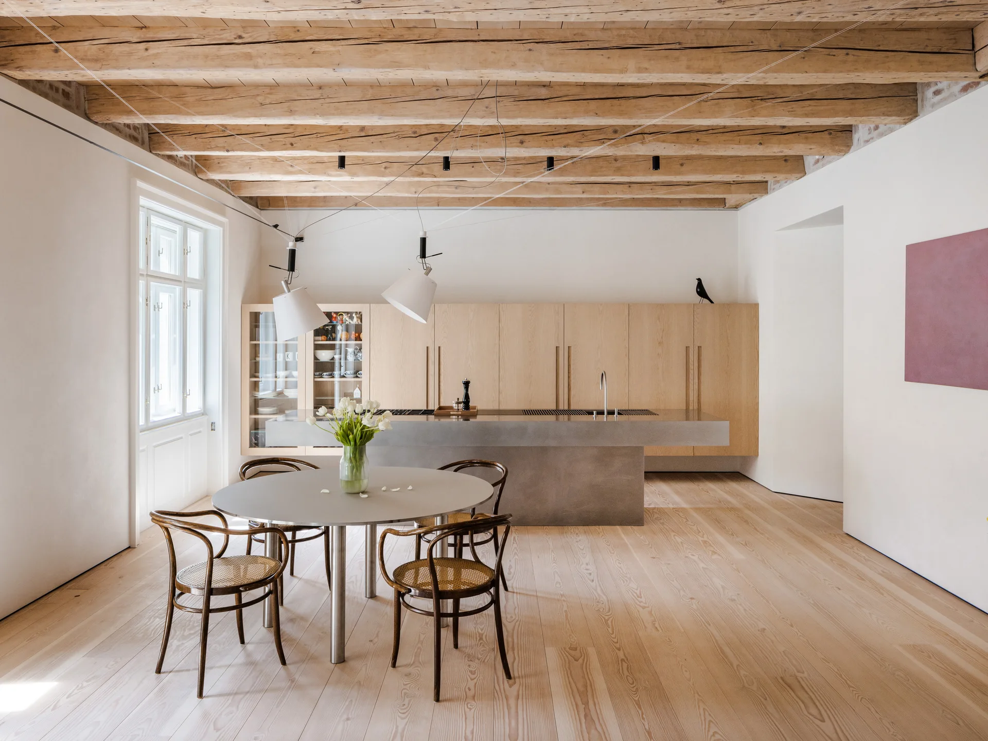 A minimalist kitchen and dining space with a round table, bentwood chairs, a concrete island, and exposed wood ceiling beams.