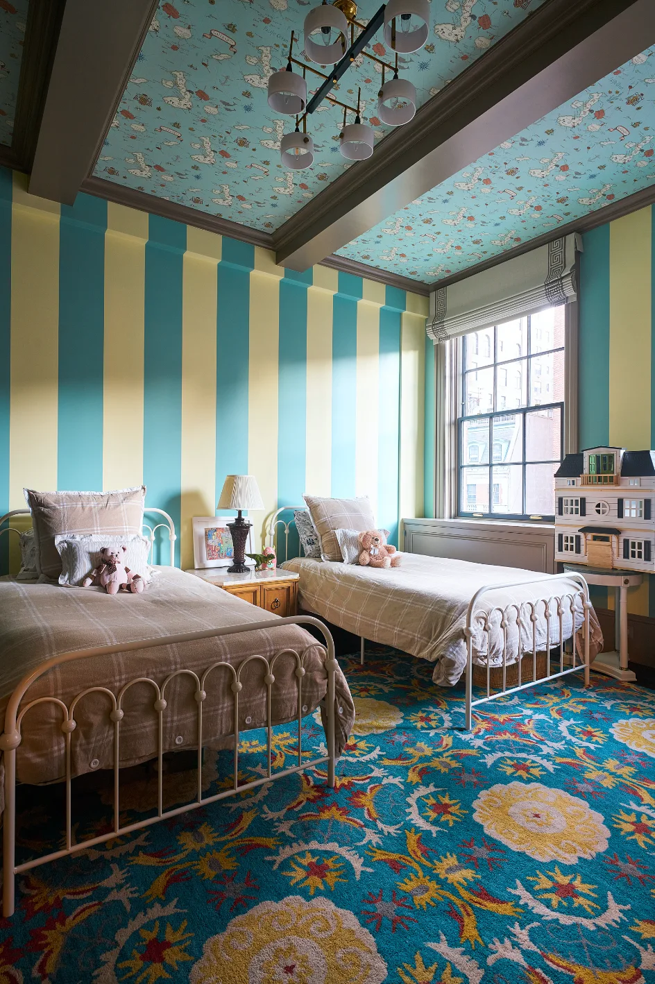 A kids' bedroom features two white metal twin beds with plaid bedding, blue and yellow striped walls, and a blue map-patterned ceiling with dark beams.