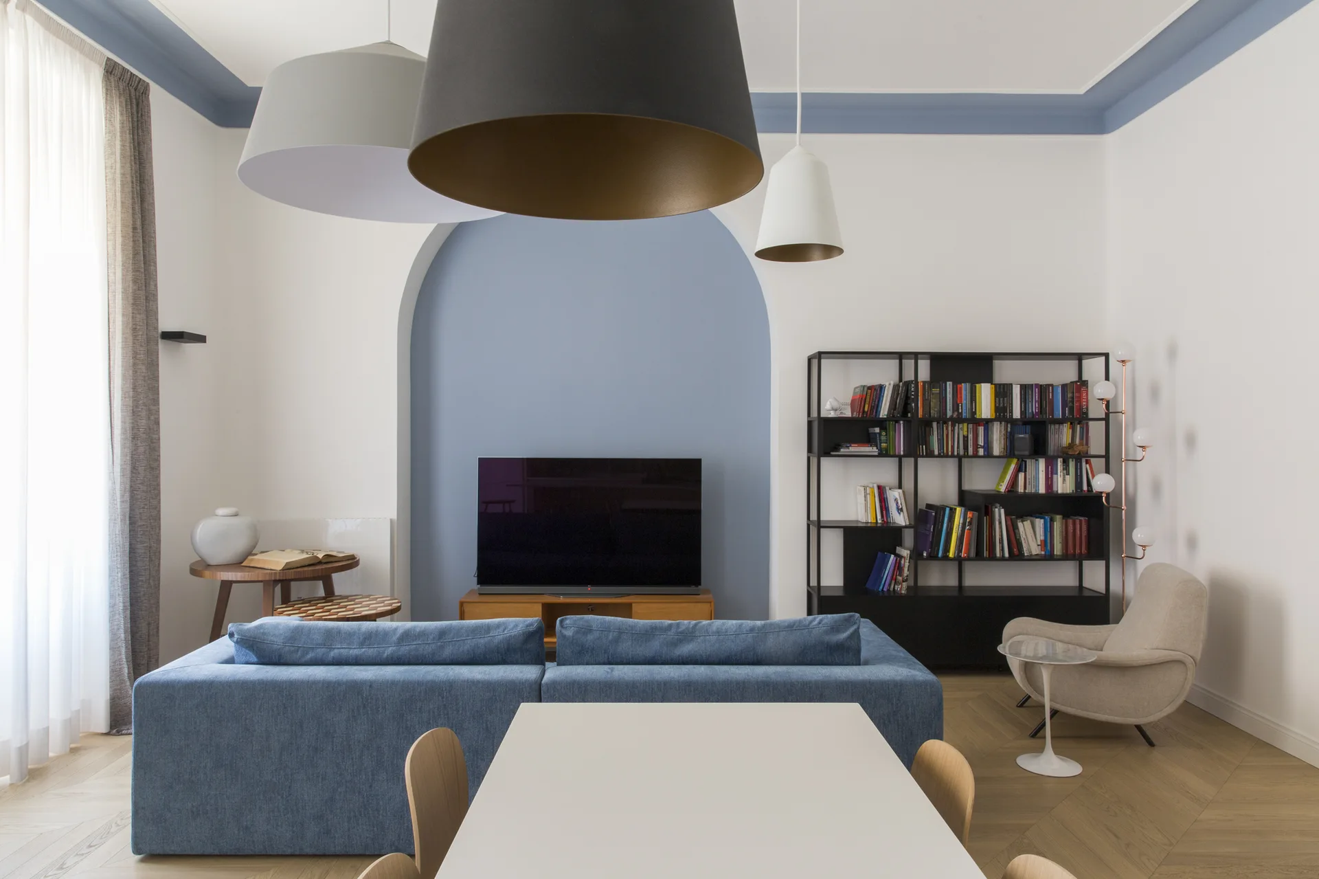 A contemporary living room featuring a blue sofa, a painted blue archway behind a television, a black bookshelf, and herringbone wood floors.