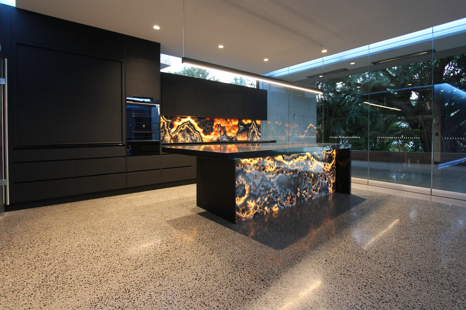 A modern kitchen features matte black cabinets, an illuminated stone island, matching backsplash, and polished concrete floors.