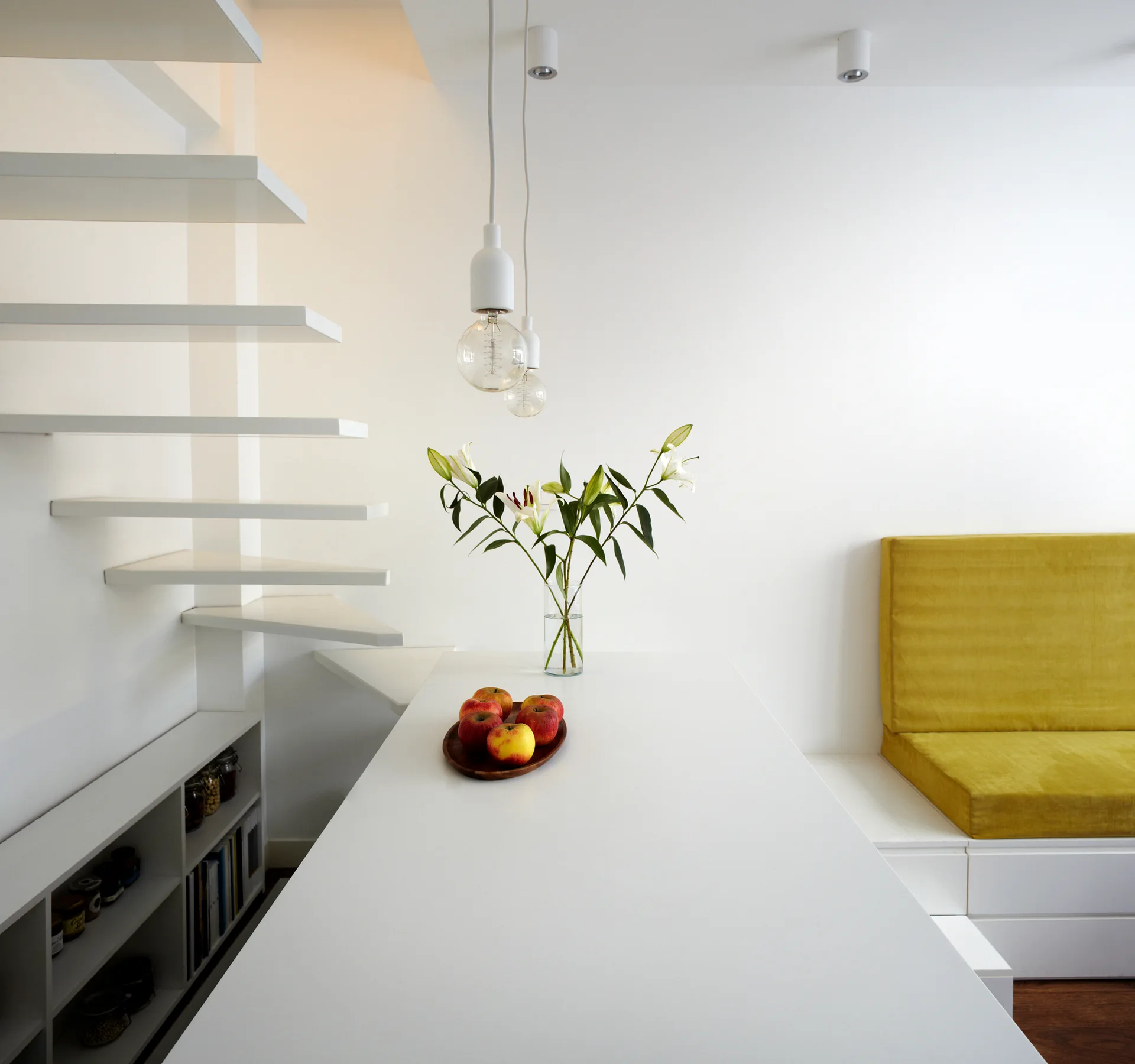 A minimalist dining room features a white table, a yellow cushioned banquette, and a white floating staircase with open risers.