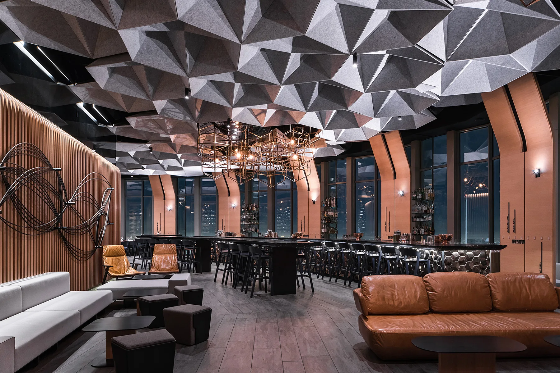 A modern lounge features a massive faceted grey ceiling, vertical wood slat walls, a long dark bar counter, and tan leather seating.