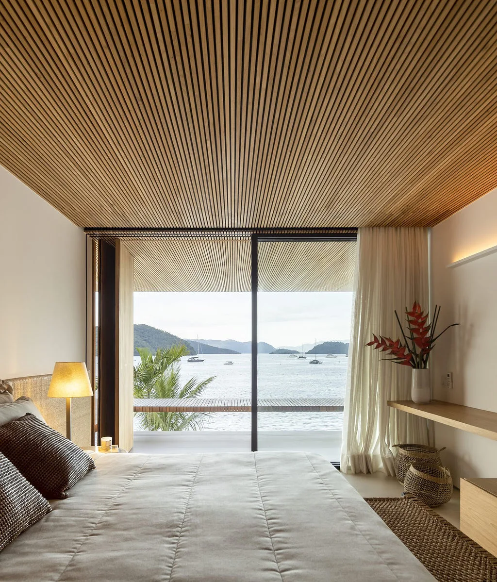 A neutral bedroom features a slatted wood ceiling, a large bed, and a sliding glass door opening to a coastal water view.