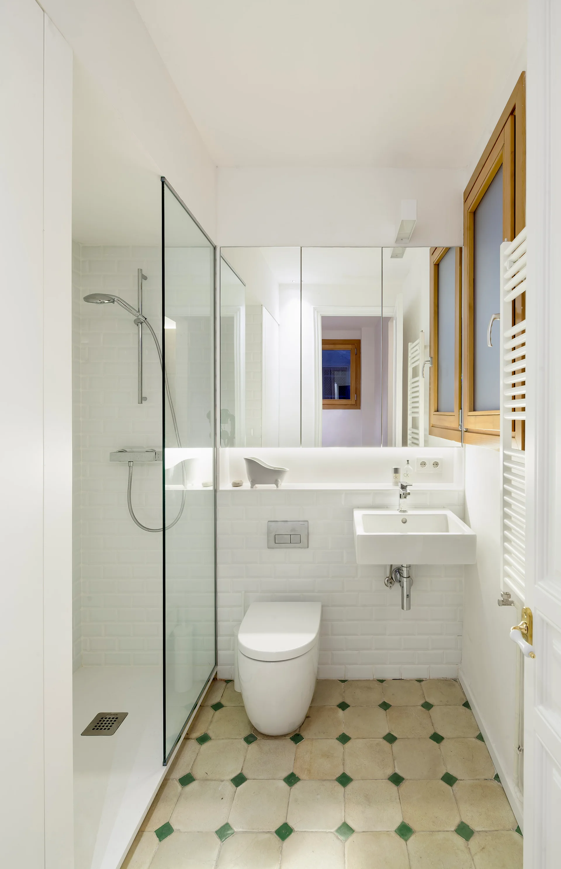 A compact white bathroom featuring a glass walk-in shower, wall-hung toilet, wall-mounted sink, and geometric floor tiles with green accents.