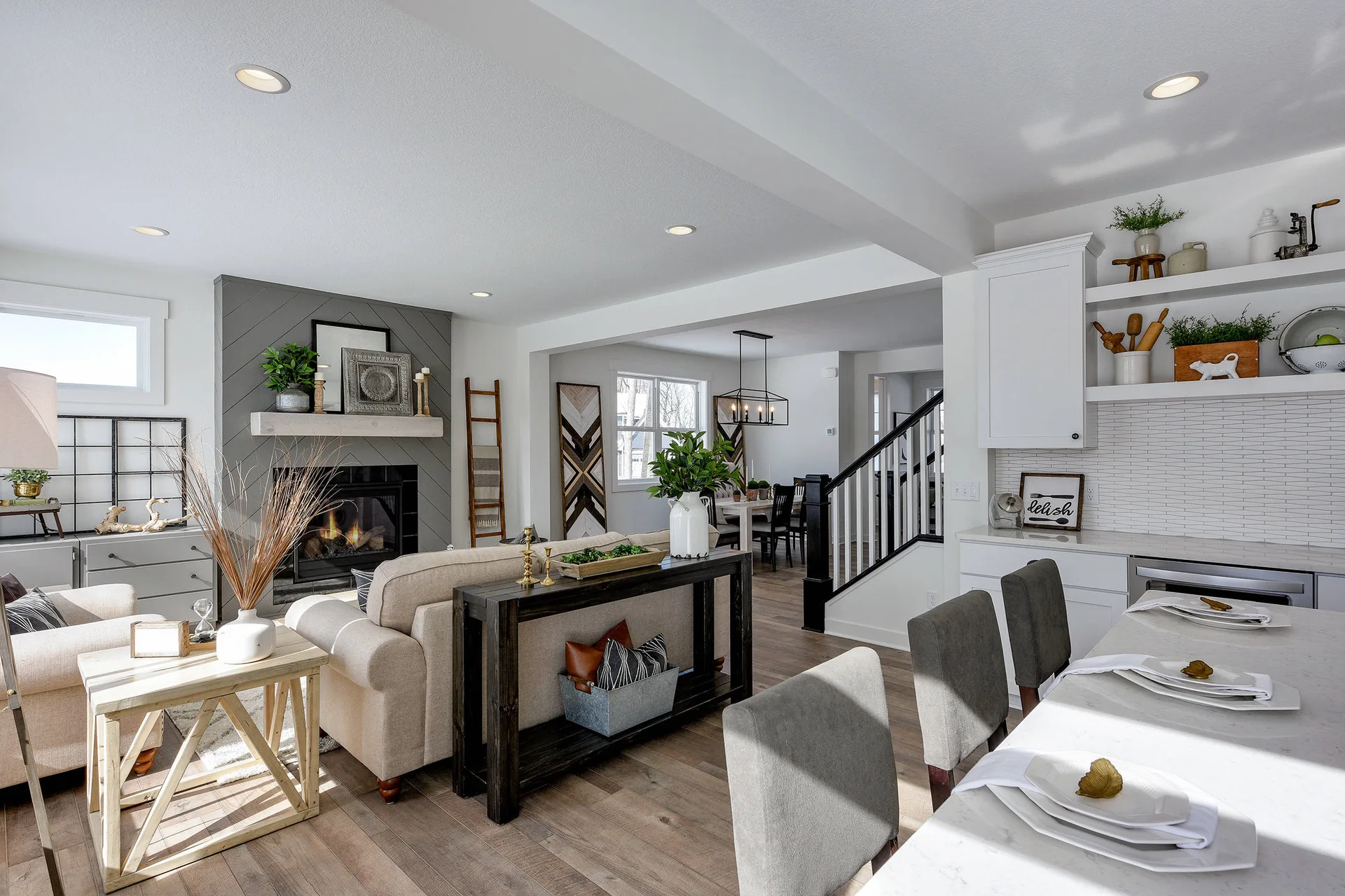 An open-concept living room features a beige sofa, a dark wood console table, and a grey fireplace with a chevron pattern next to a kitchen.