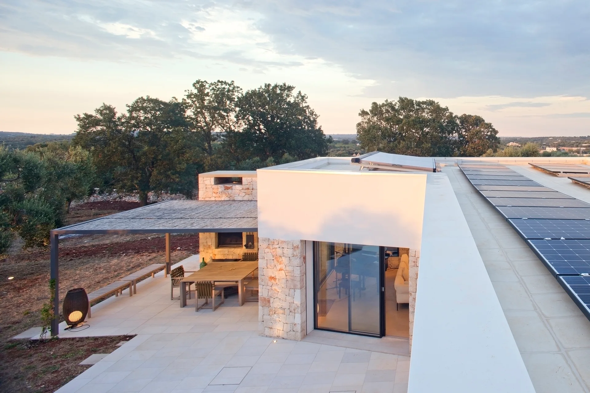 An outdoor patio features a wooden dining set under a slatted pergola next to a modern stone-and-stucco home with solar panels on the flat roof.
