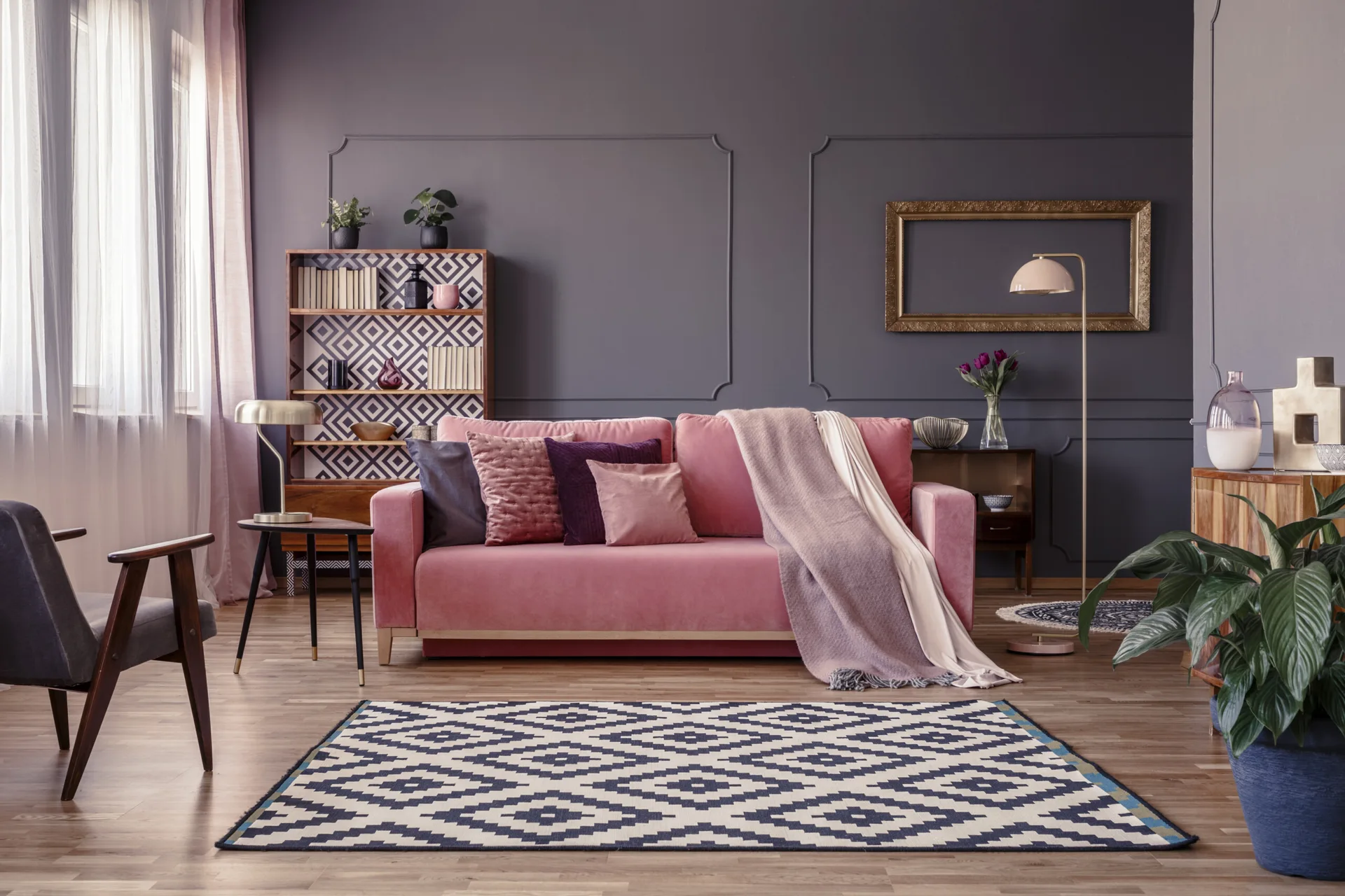 A pink velvet sofa sits in front of dark grey paneled walls next to a wooden bookshelf and a geometric patterned rug.