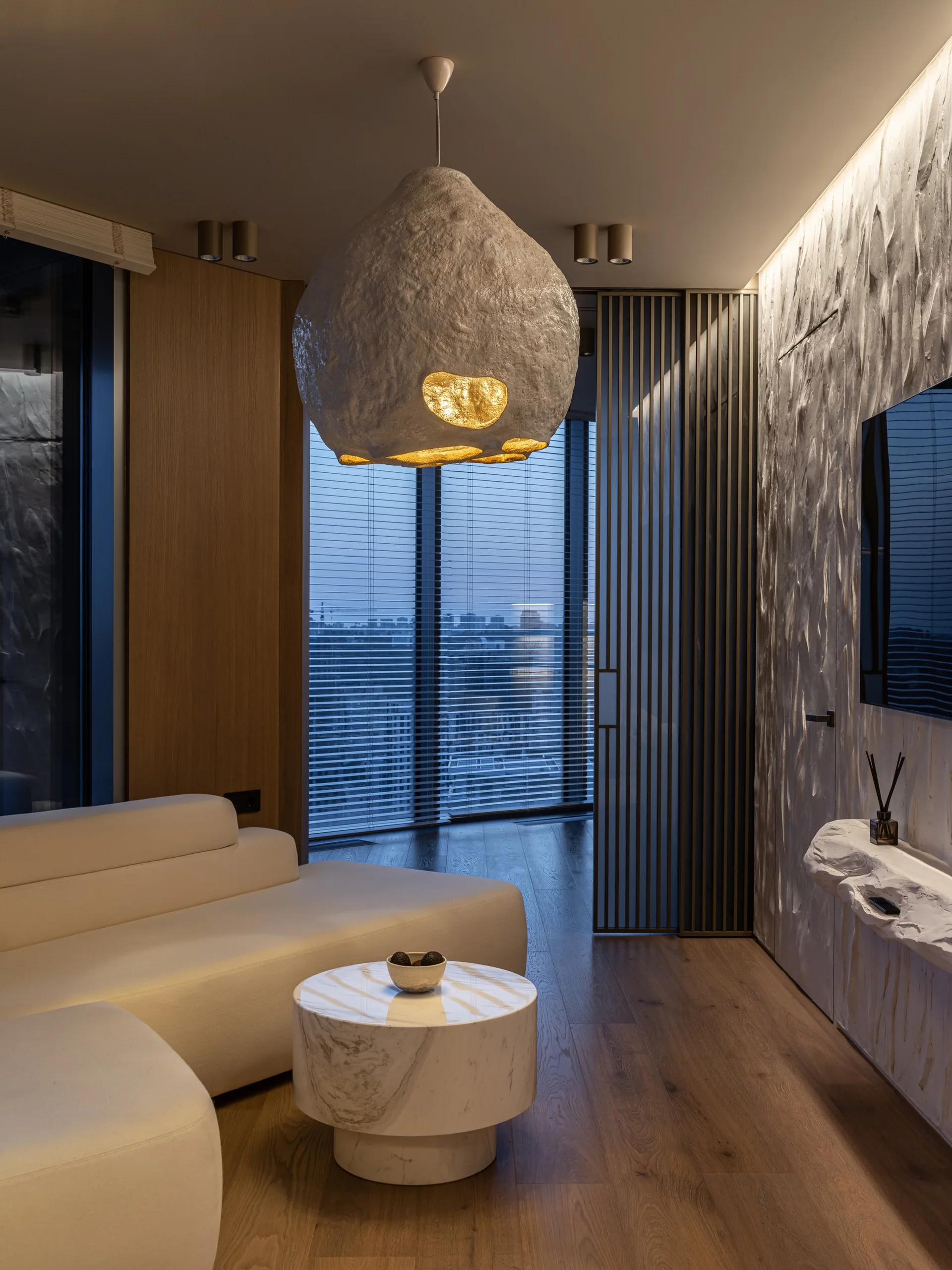 A modern living room features a white sectional sofa, a marble coffee table, a large textured pendant light, and a rough white feature wall.