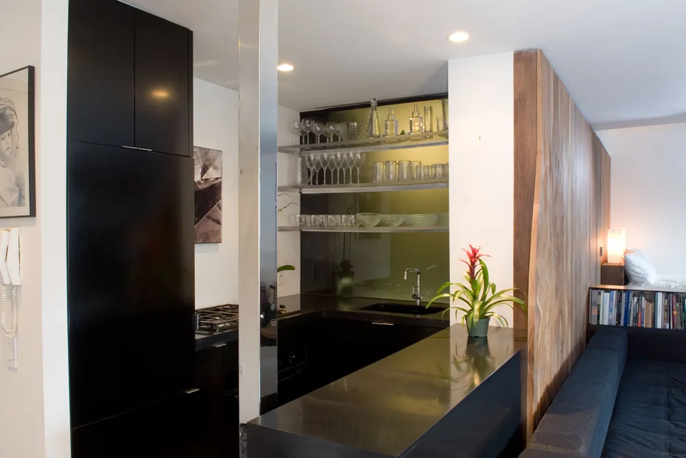A modern compact kitchen featuring black matte cabinetry, a dark stone countertop, open glass shelving, and a vertical wood partition wall.